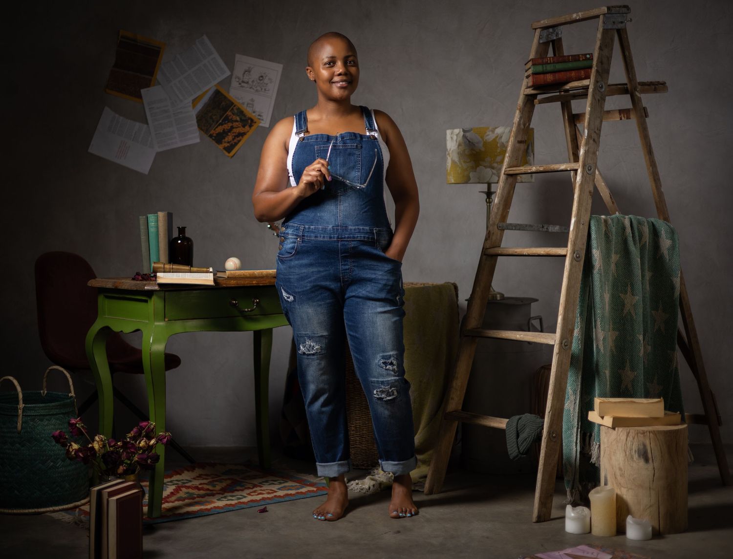 Woman in denim overalls stands confidently in an artistic studio space with a wooden ladder and green desk.
