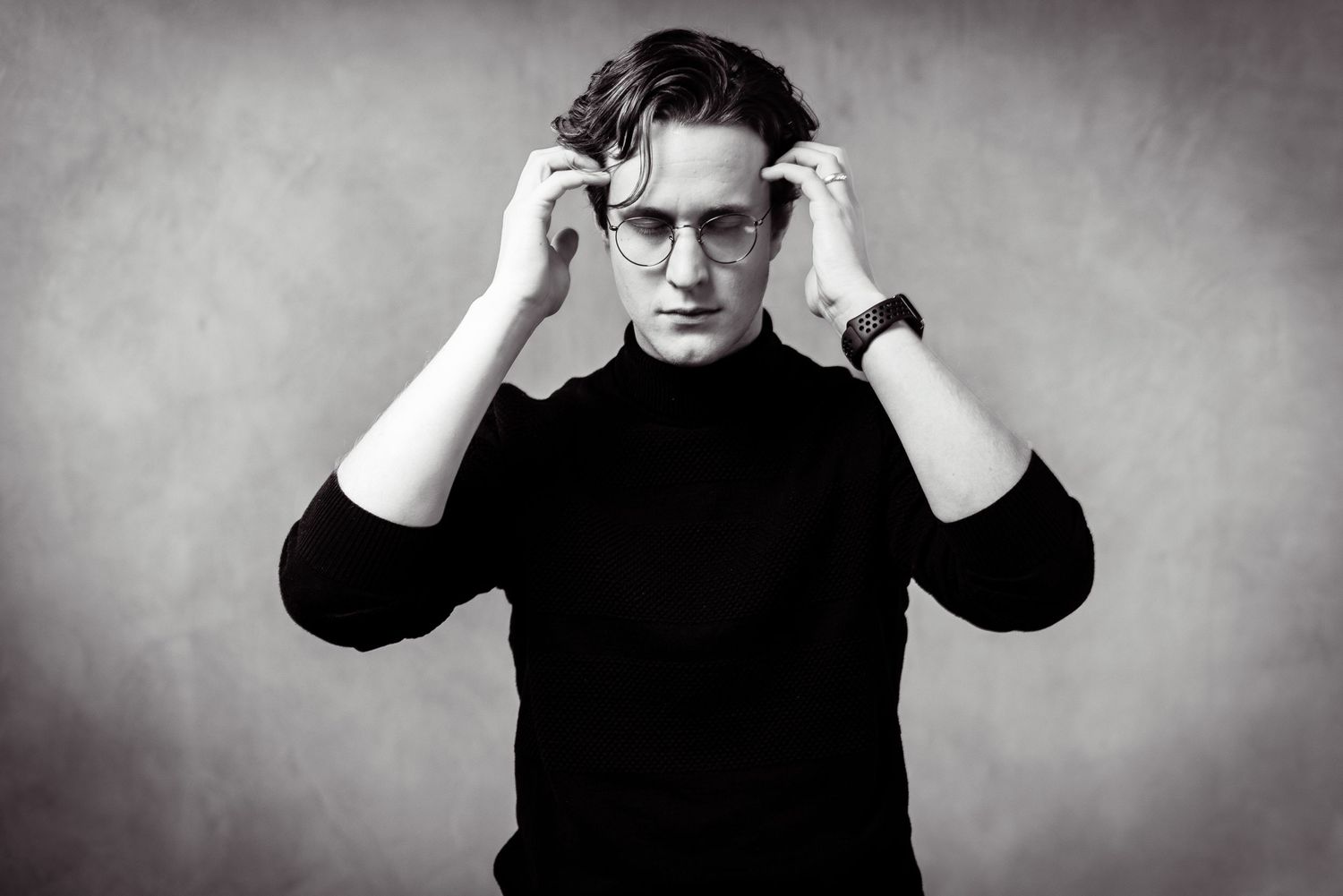 A black and white portrait of someone in a dark turtleneck sweater adjusting their glasses against a textured backdrop.