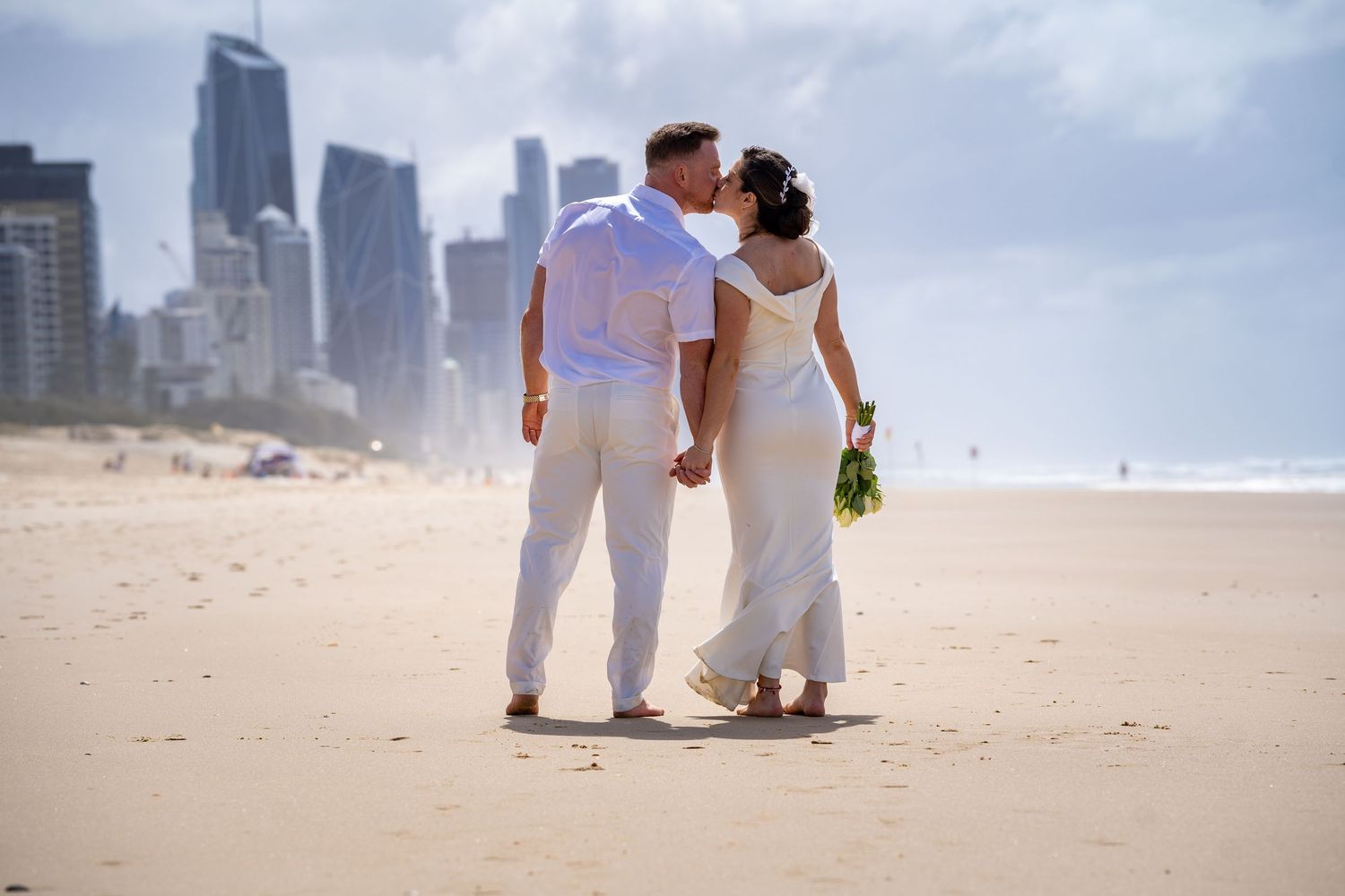 A couple in white attire walking together on a beach with city skyscrapers in the background.