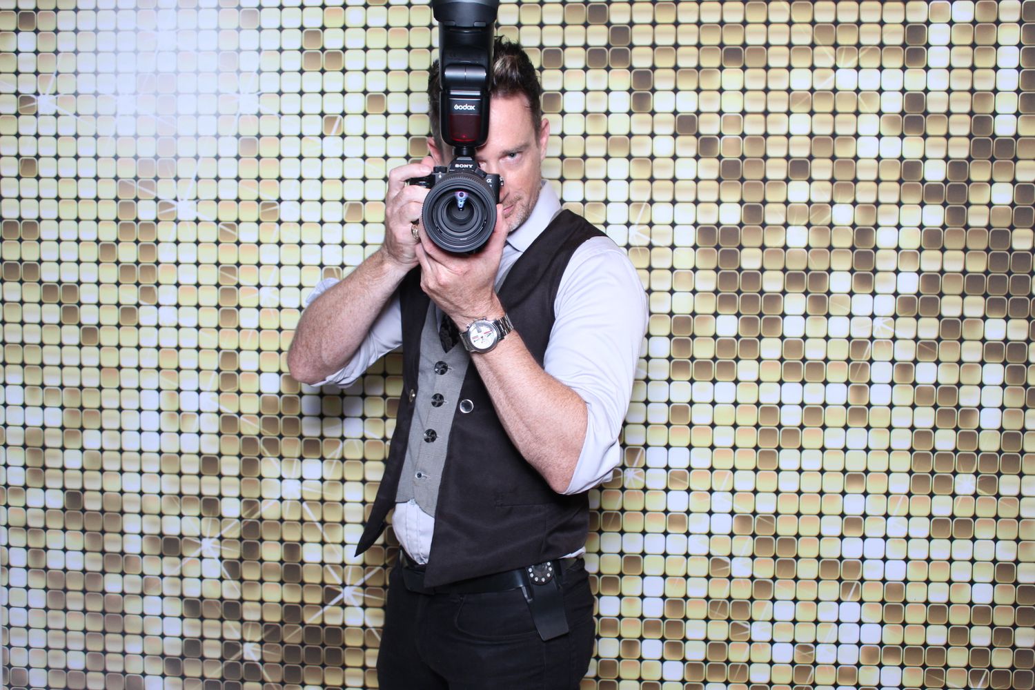 Professional photographer wearing black vest holds camera against sparkly gold sequin backdrop.