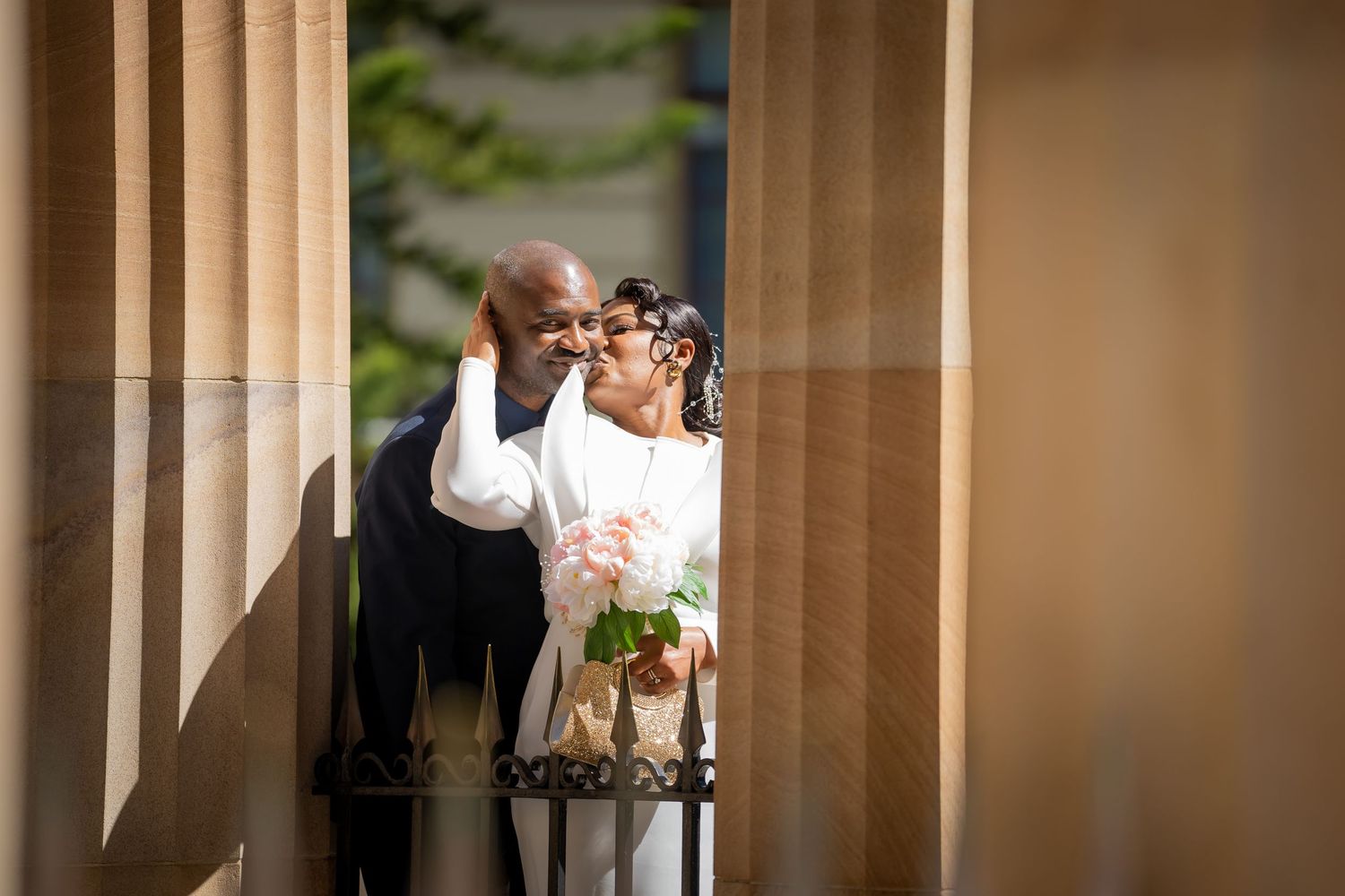 A couple in formal wedding attire shares an intimate moment between elegant stone pillars at an outdoor venue.