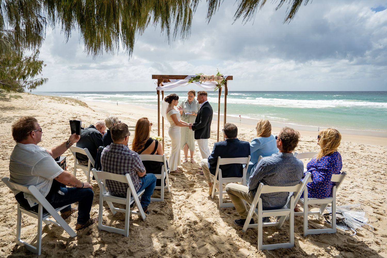 Guests seated in white chairs on a tropical beach during a destination wedding ceremony with ocean views.