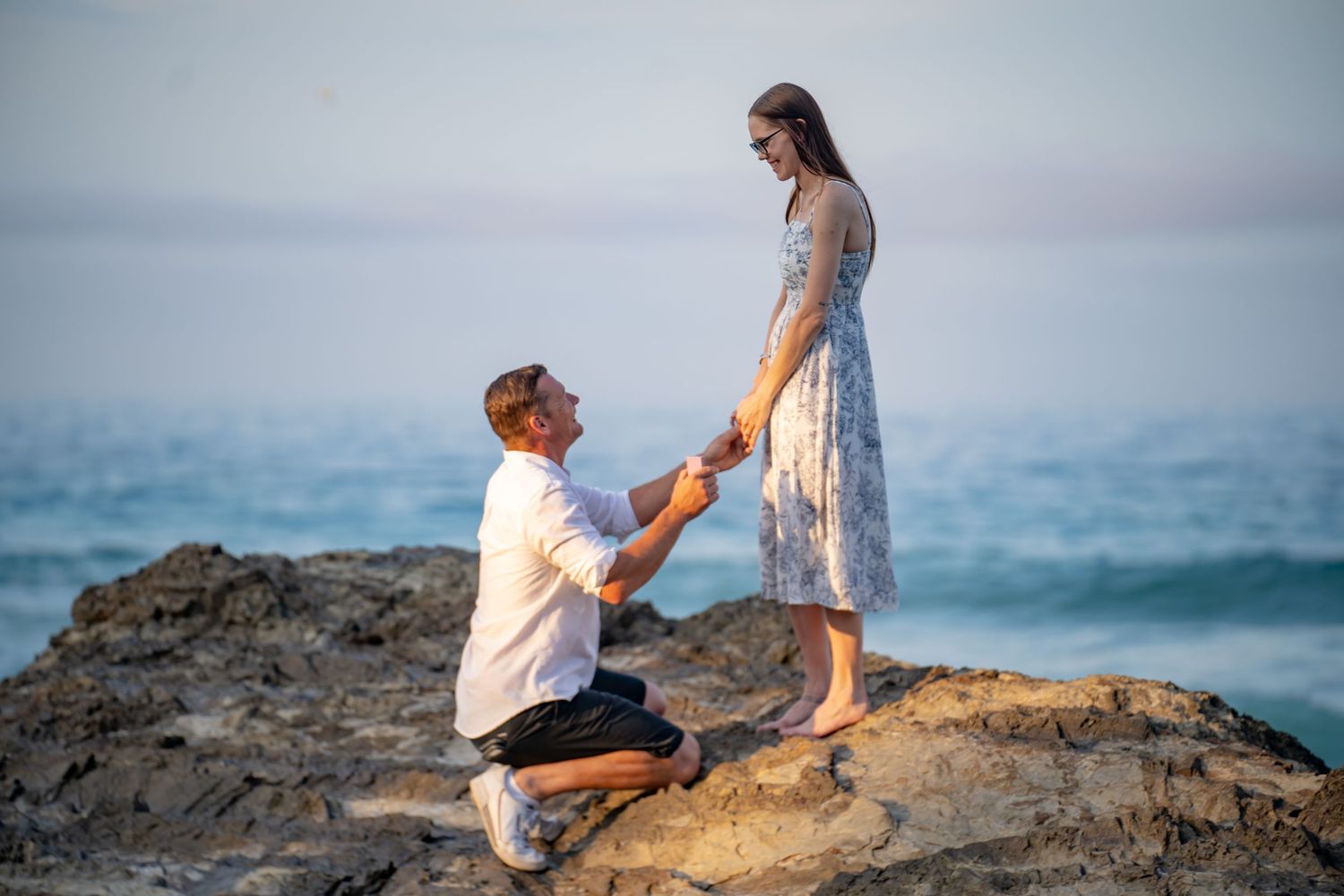 A couple shares a romantic moment on rocky coastal cliffs during sunset with ocean waves in the background.
