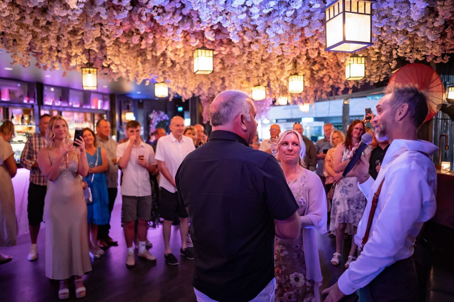 Wedding guests dance under a canopy of cherry blossoms and glowing lanterns during an elegant evening reception.
