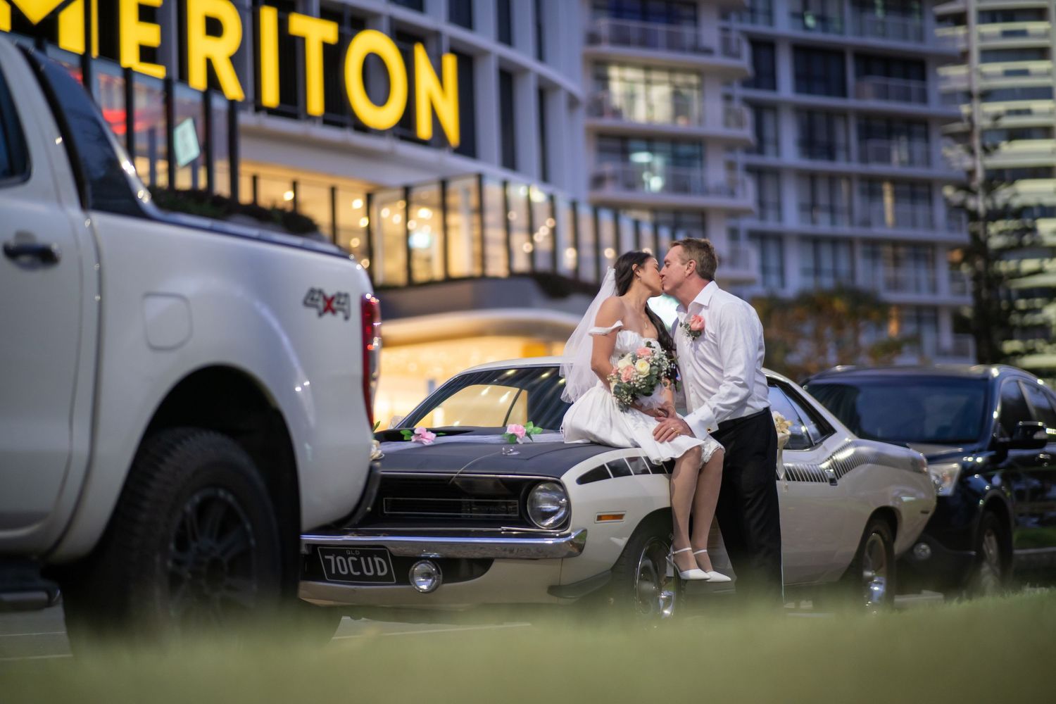 A group of people gather outside the Meriton hotel building at night with cars parked nearby.