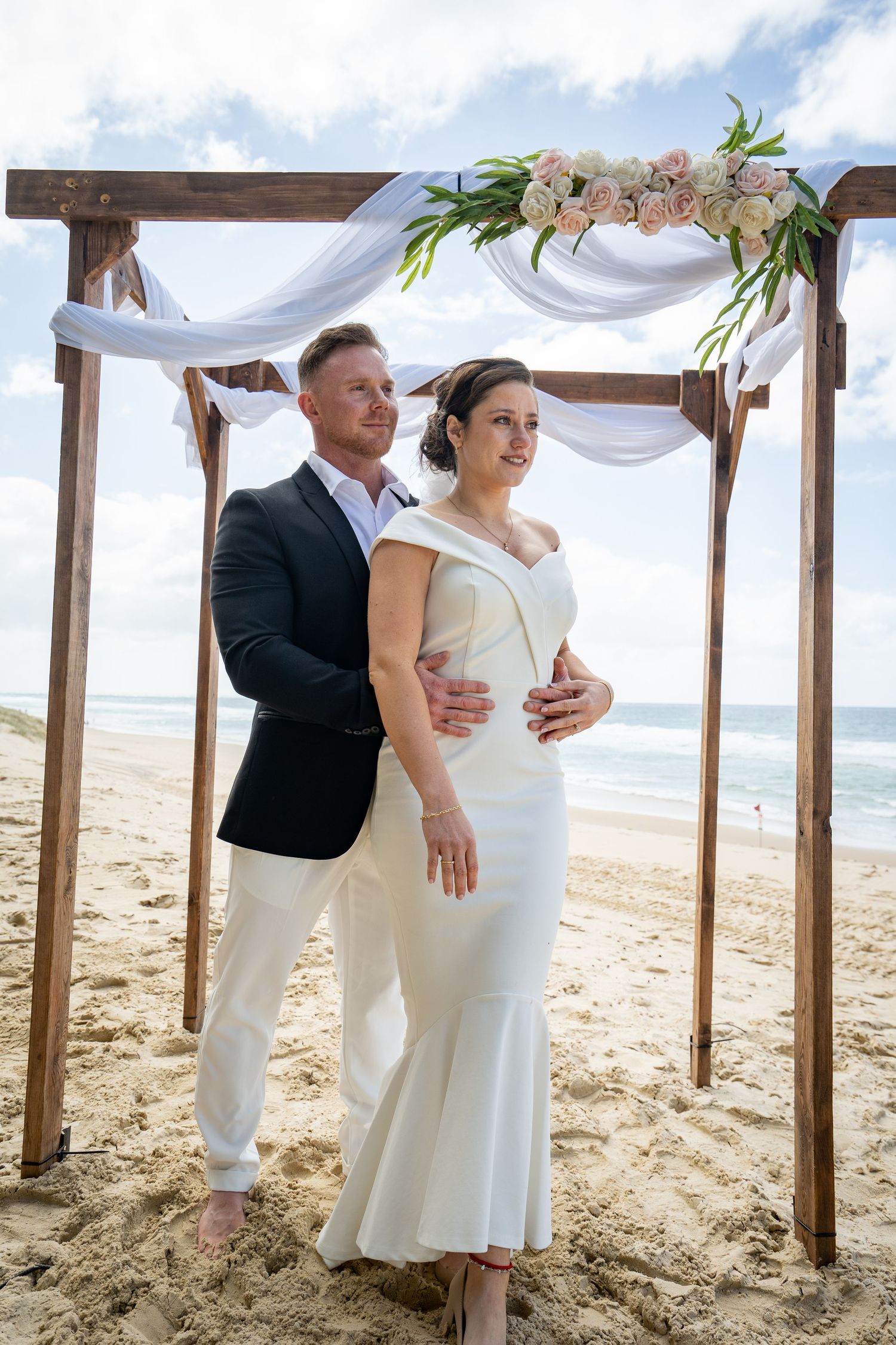 A beachside wedding ceremony setup with wooden posts draped in white fabric and flowers along a sandy shoreline.