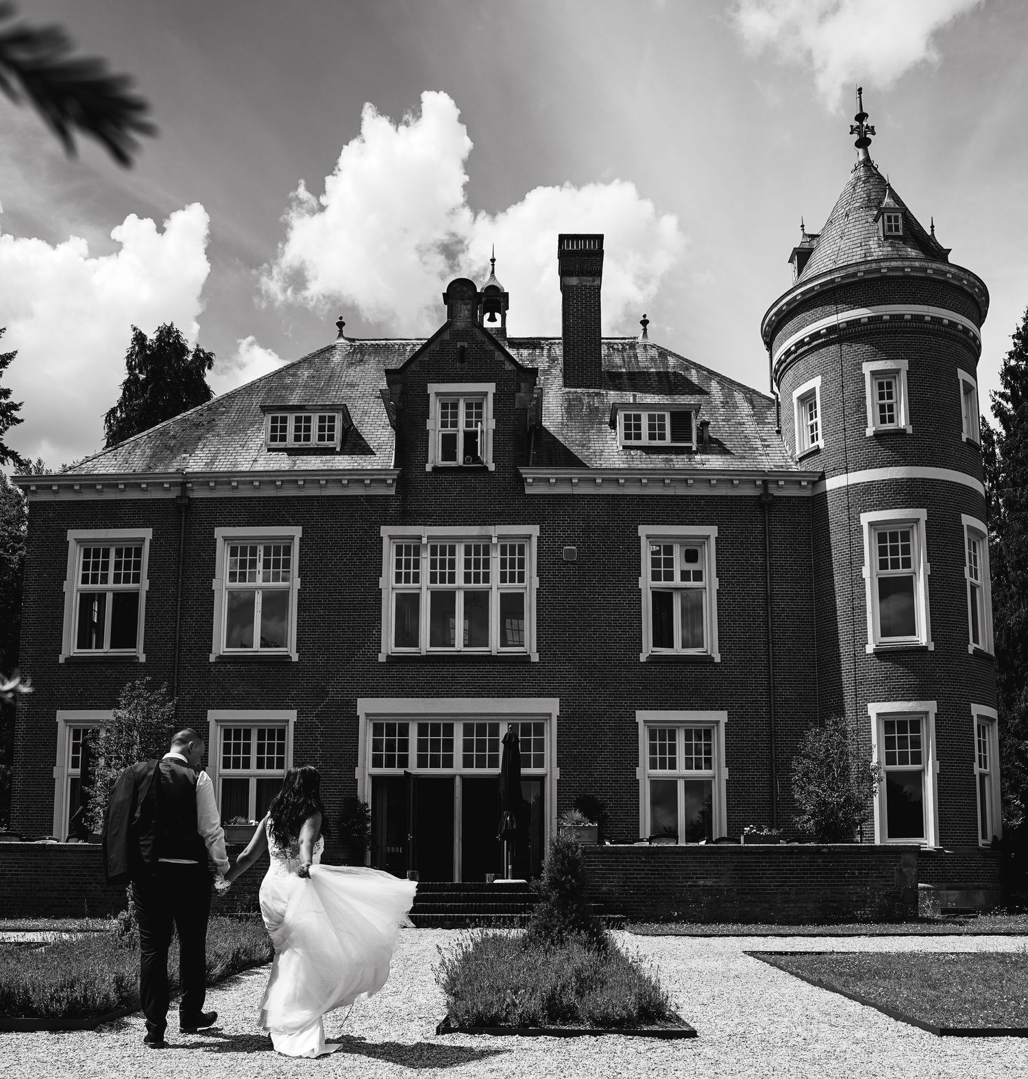 Bruid en bruidegom wandelen hand in hand naar een groot kasteel in zwart-wit sfeer.