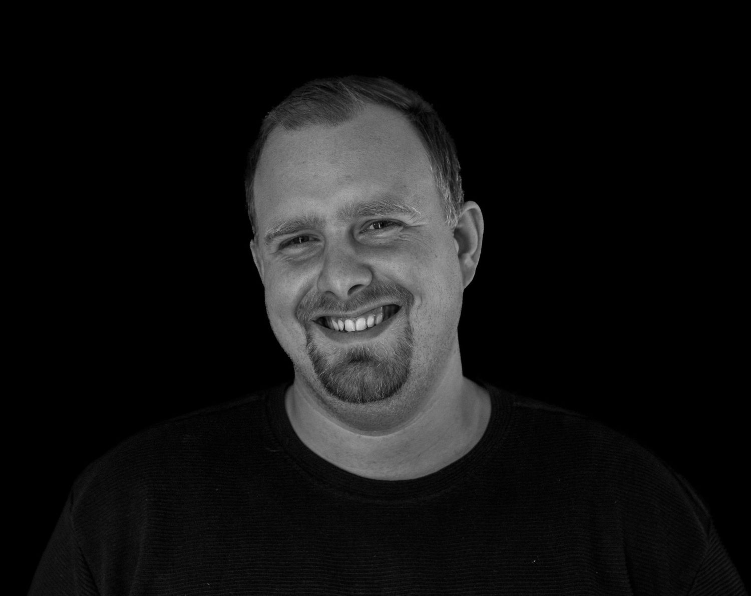 Man in zwart shirt lacht naar de camera, portret op zwarte achtergrond.