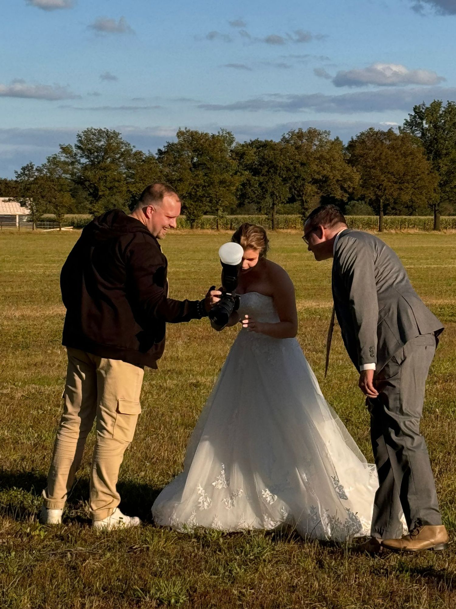 Bruidspaar bekijkt trouwfoto’s met hun fotograaf in een zonnige buitenlocatie.