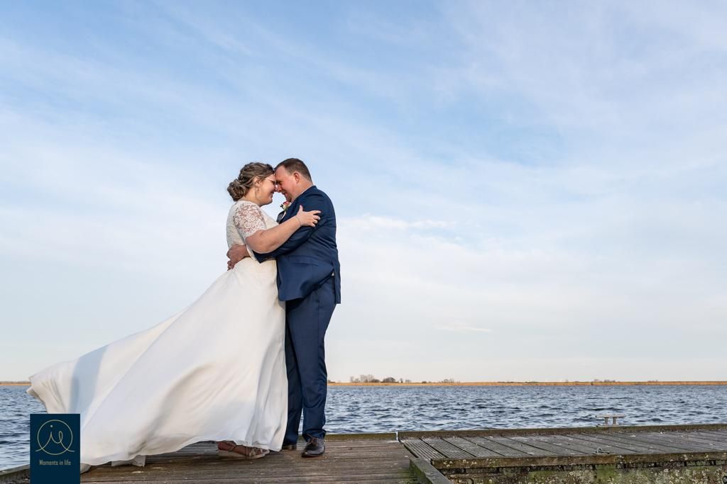 Bruidspaar omhelst elkaar liefdevol op een steiger bij het water onder een blauwe lucht.