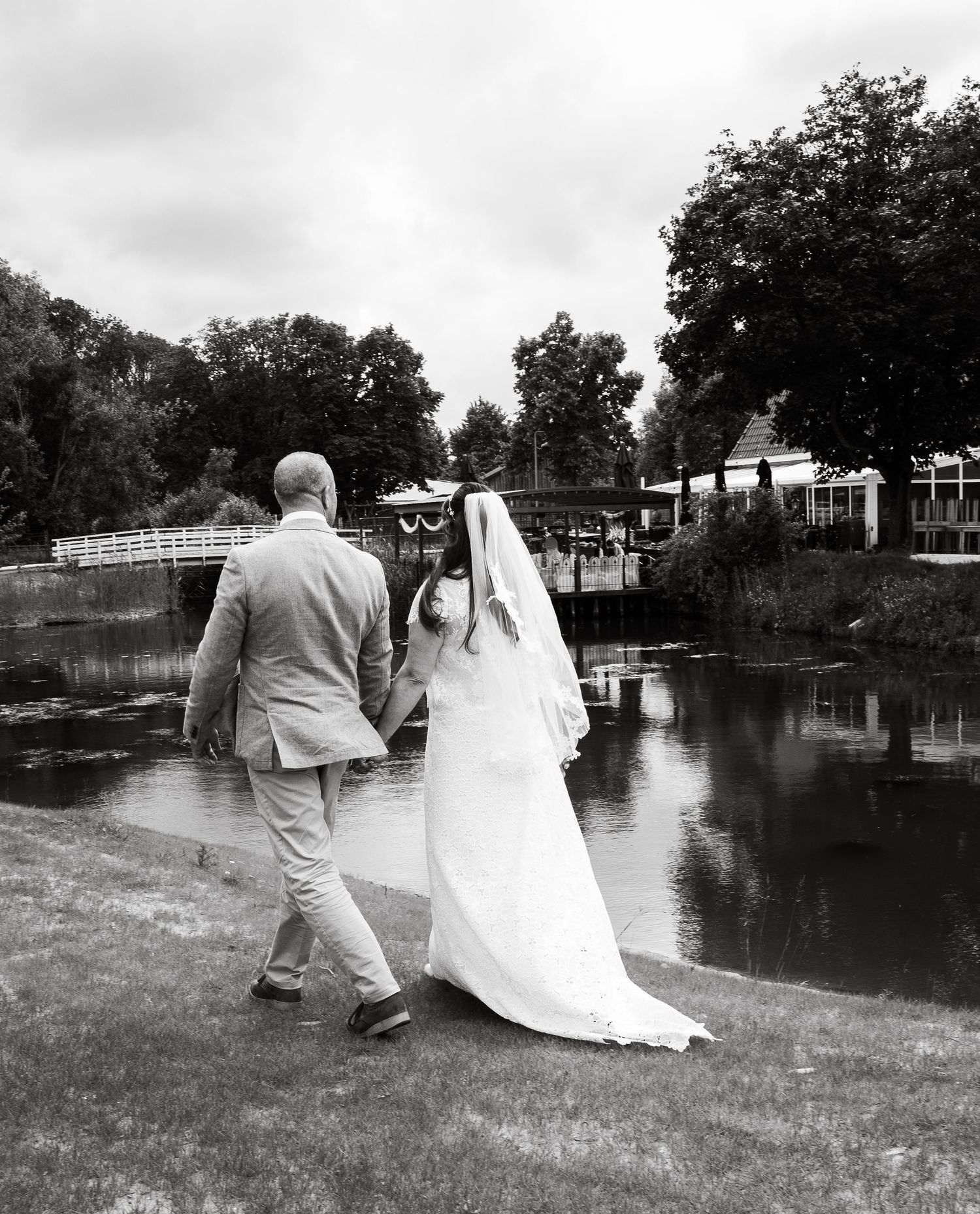 Bruid en bruidegom lopen hand in hand langs een vijver in een rustige parksetting.
