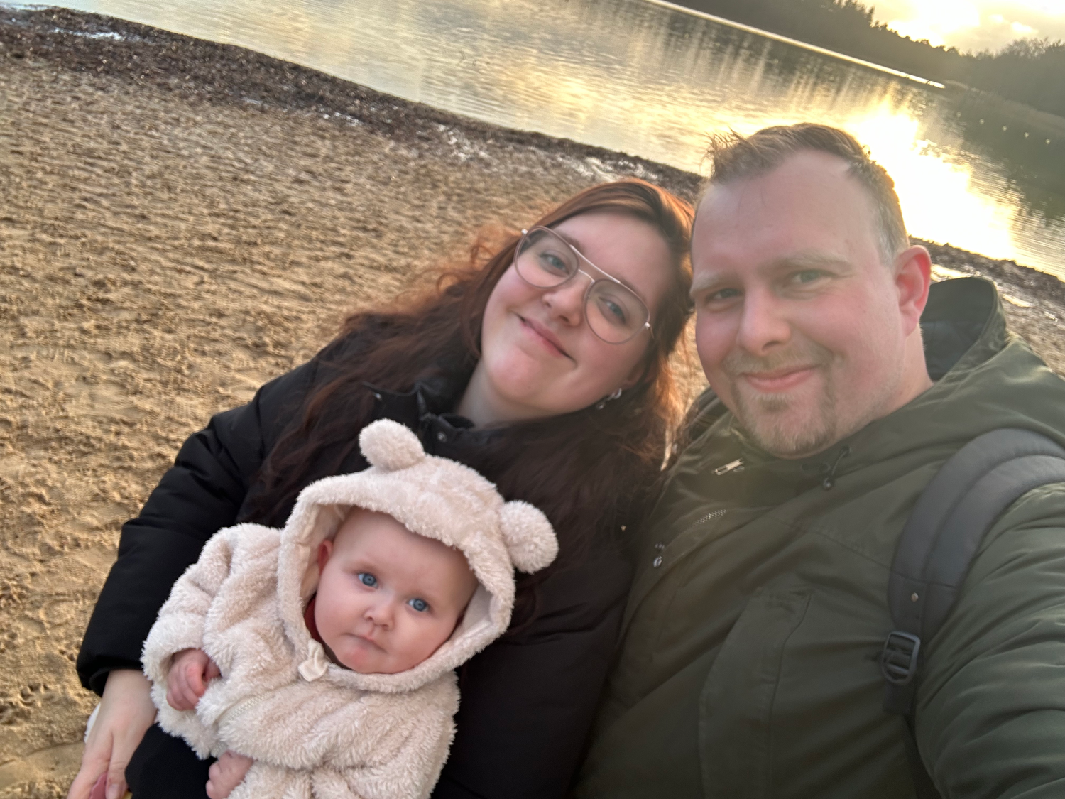 Stel met baby aan het meer bij zonsondergang, glimlachend in de camera.