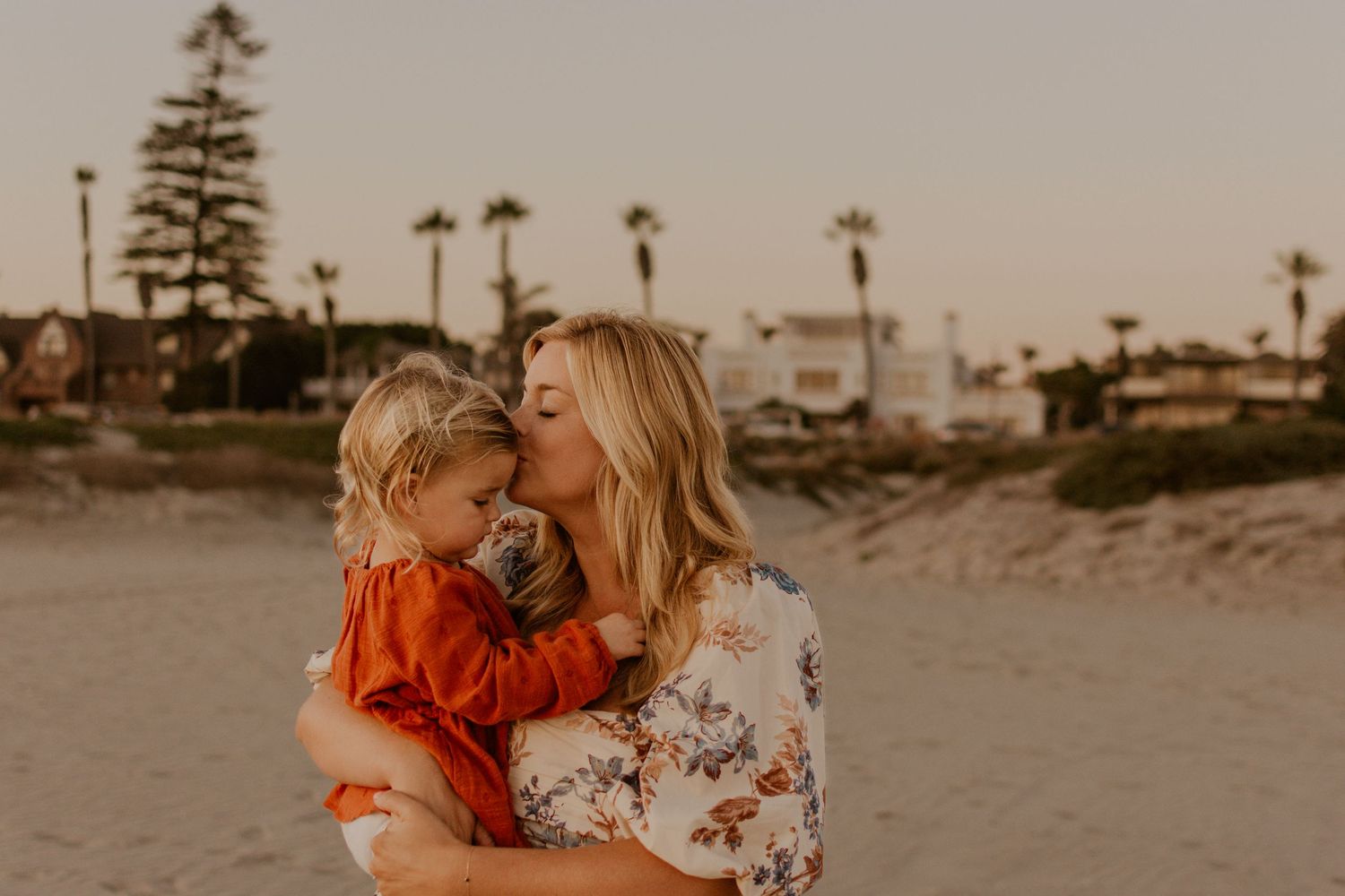 Coastal family portrait at golden hour showing a mother and child in a floral dress sharing a heartfelt embrace on the beach.