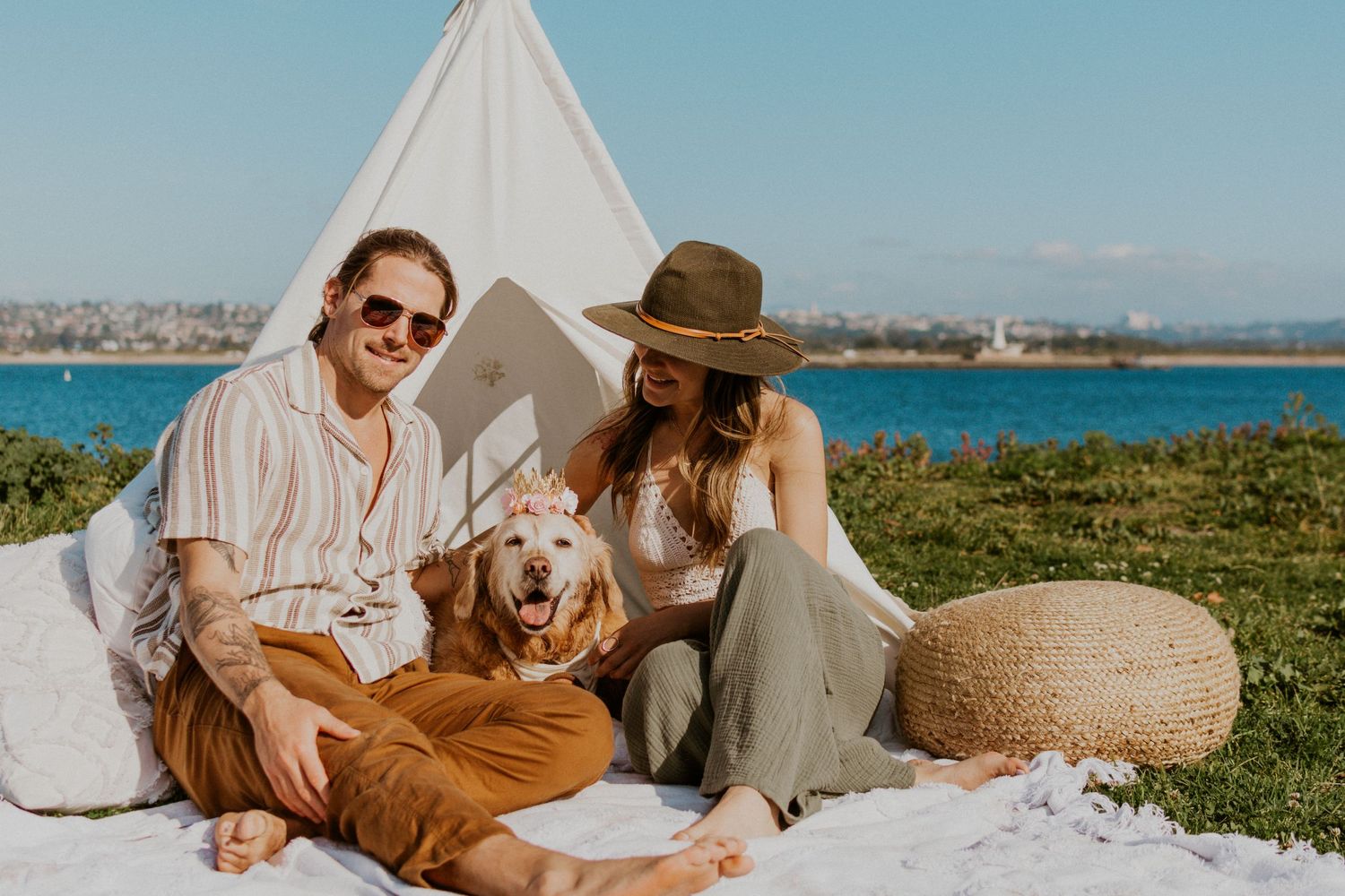 Pet and lifestyle session with couple and golden retriever relaxing under a white teepee setup on a beachside blanket.