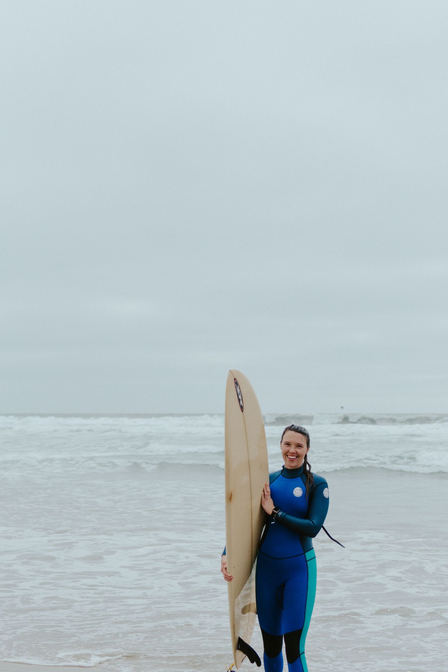 Lifestyle session on a foggy beach with surfer walking along the shoreline in natural light.