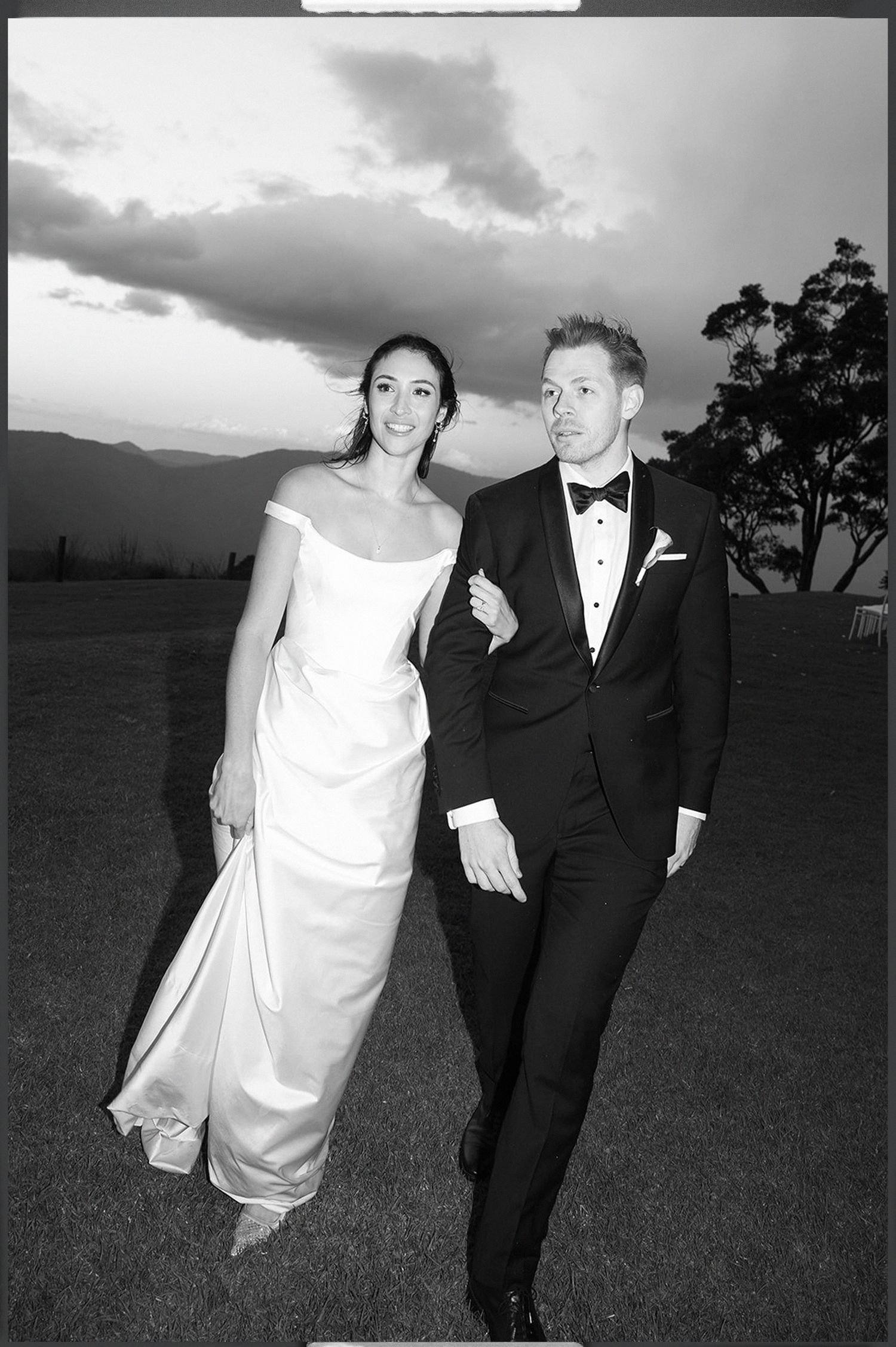 A couple in wedding attire runs playfully across a grassy field at sunset with dramatic clouds in the background.