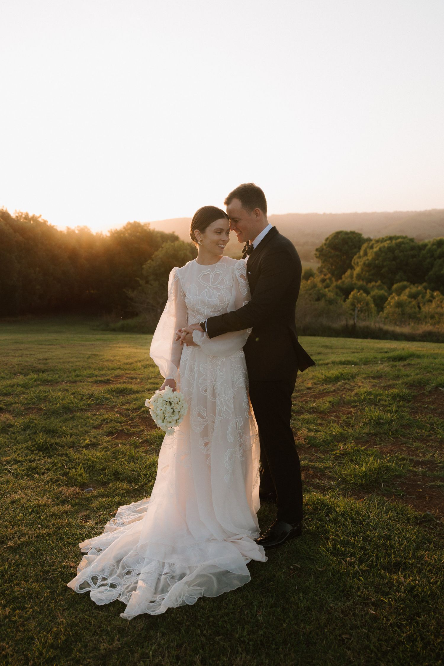 A couple shares an intimate sunset moment in a grassy field wearing wedding attire during golden hour.