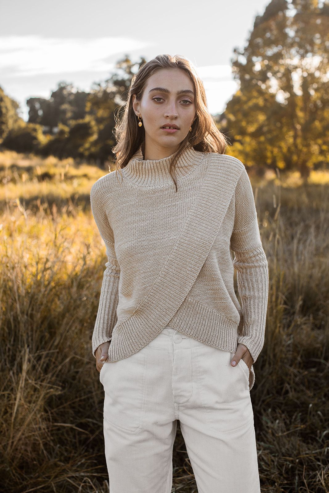 Model wearing beige knit sweater and white pants poses in golden autumn field during sunset.