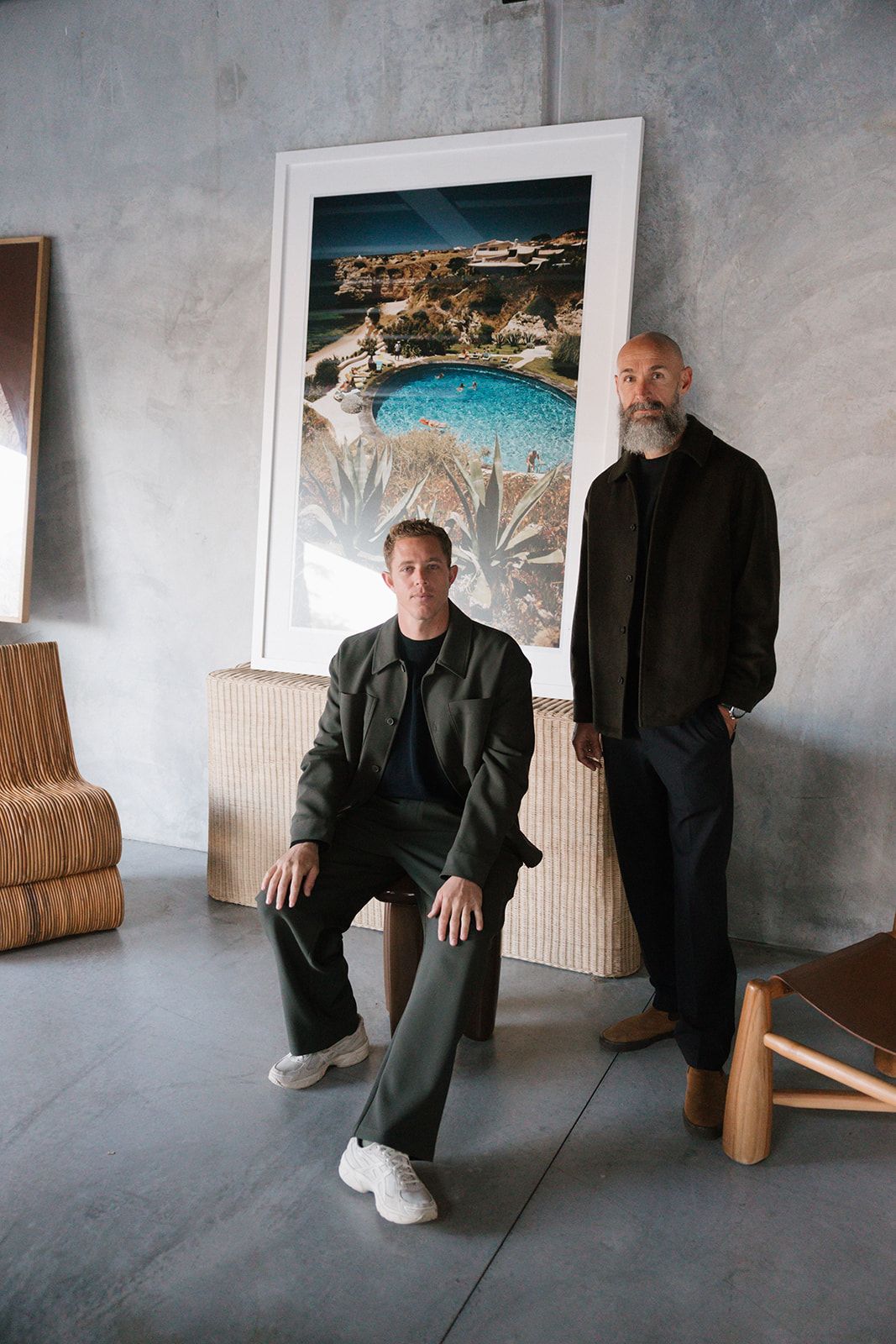 Two figures in black attire sit and stand in a minimalist room with framed artwork on the walls.