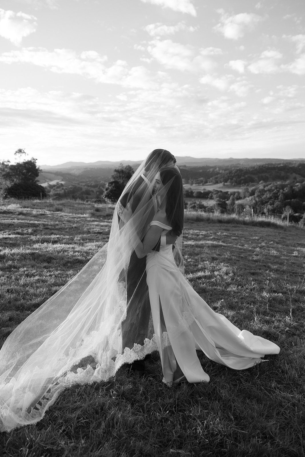Black and white photo of a bride in a flowing lace veil embracing her partner on a grassy hilltop overlooking rolling countryside.