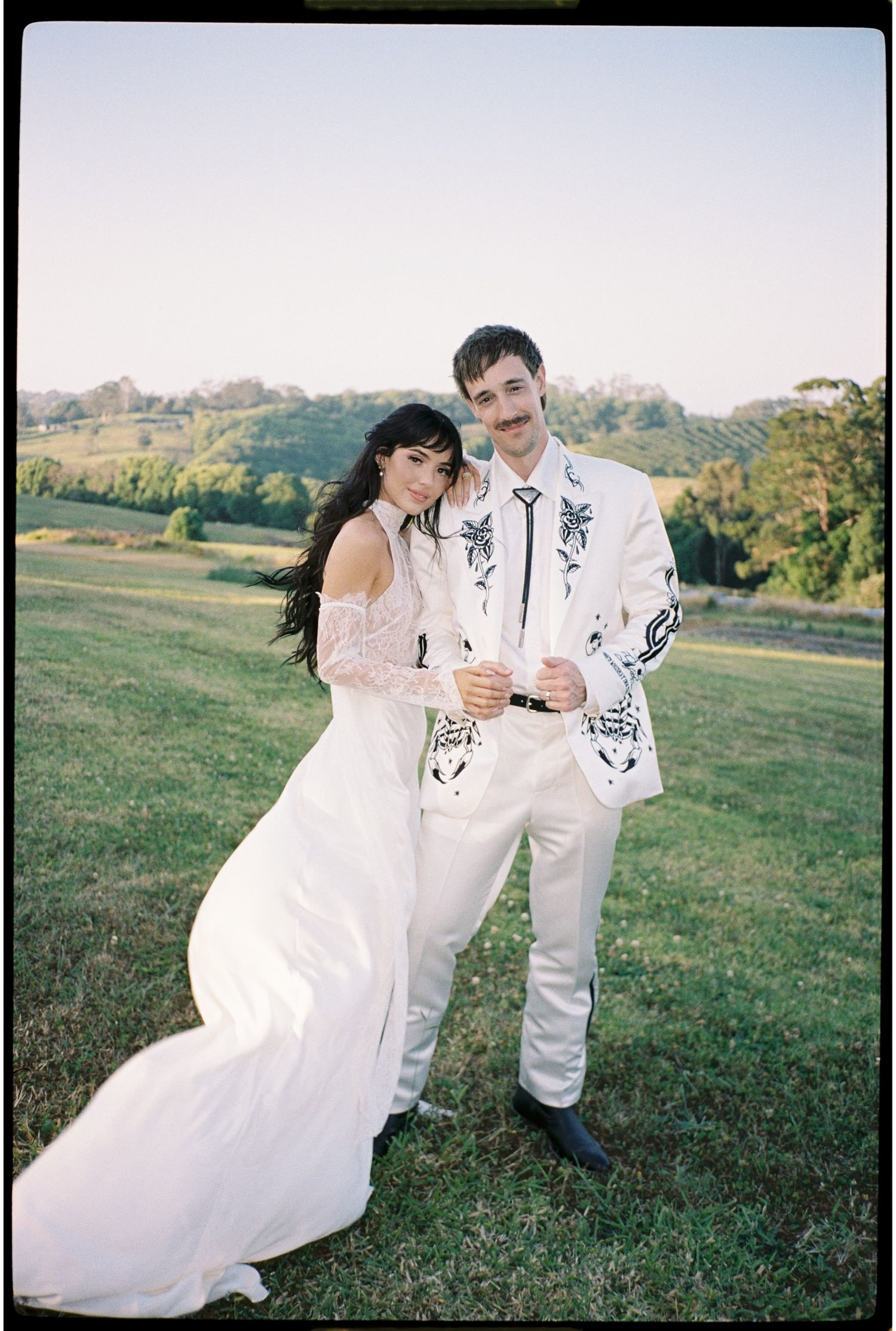 Bride in white gown and groom in embroidered white suit pose together in a lush green field at golden hour.