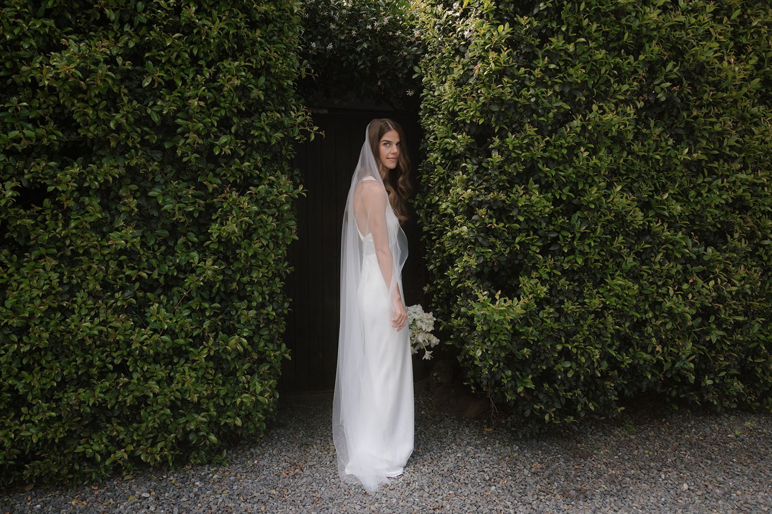 A person in a white wedding dress stands elegantly against a lush green hedge backdrop in a garden setting.