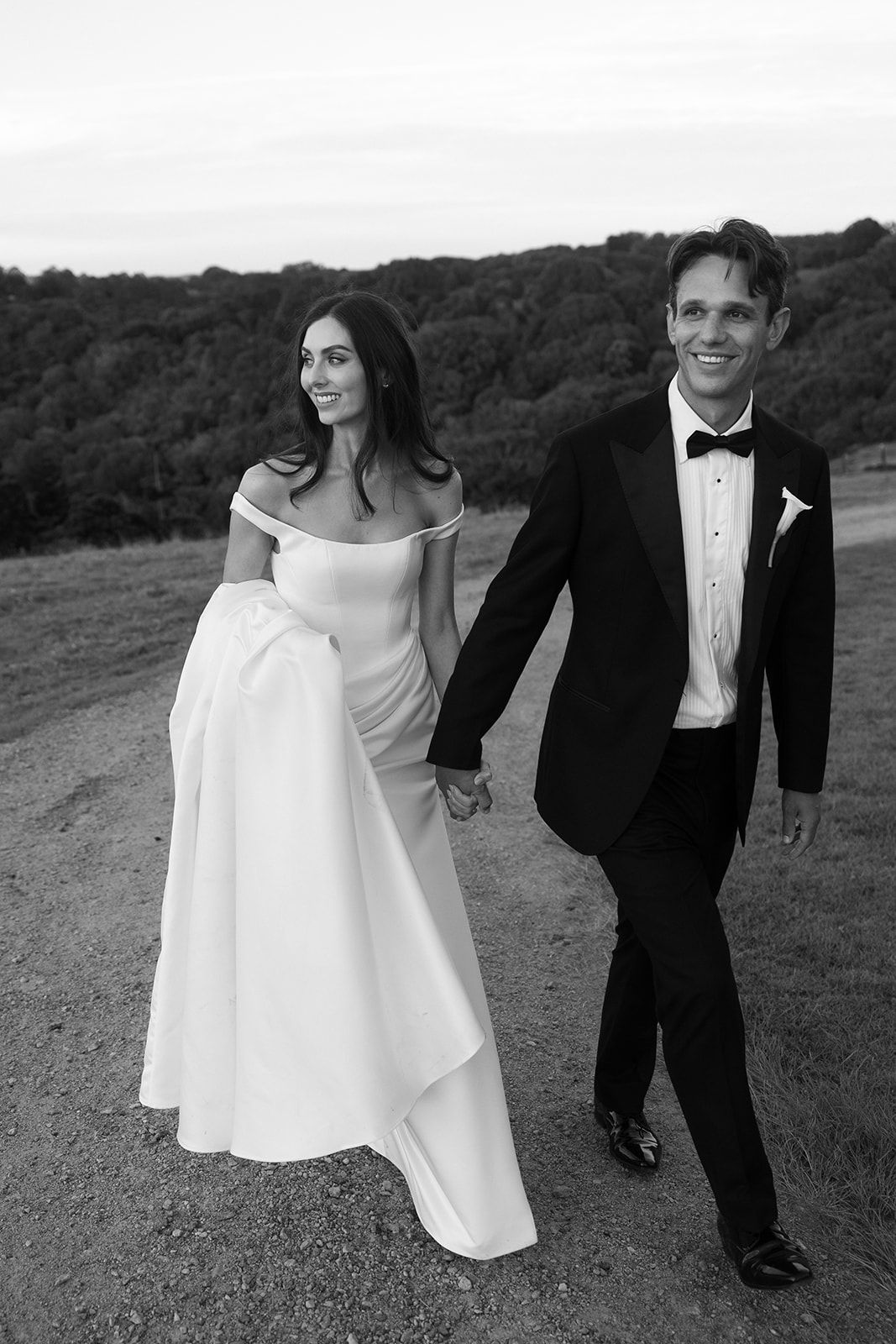 Black and white photo of a smiling bride and groom holding hands walking outdoors on their wedding day.