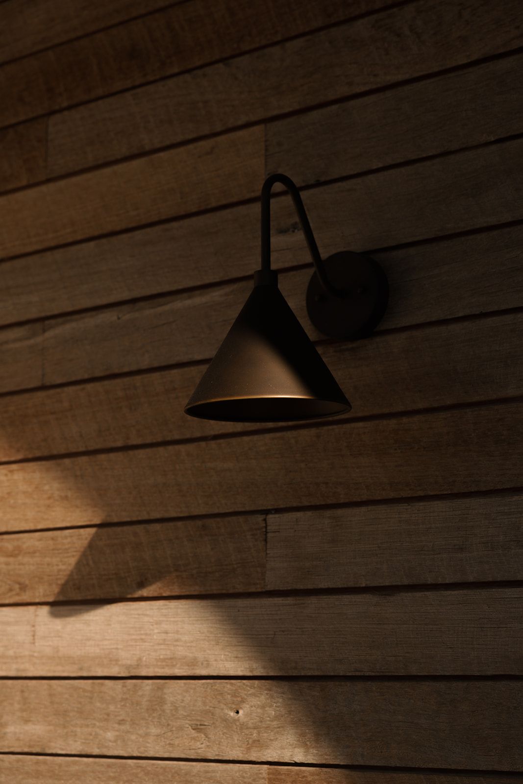 Black cone-shaped wall sconce mounted on rustic wooden plank wall, casting dramatic shadows in warm ambient light.