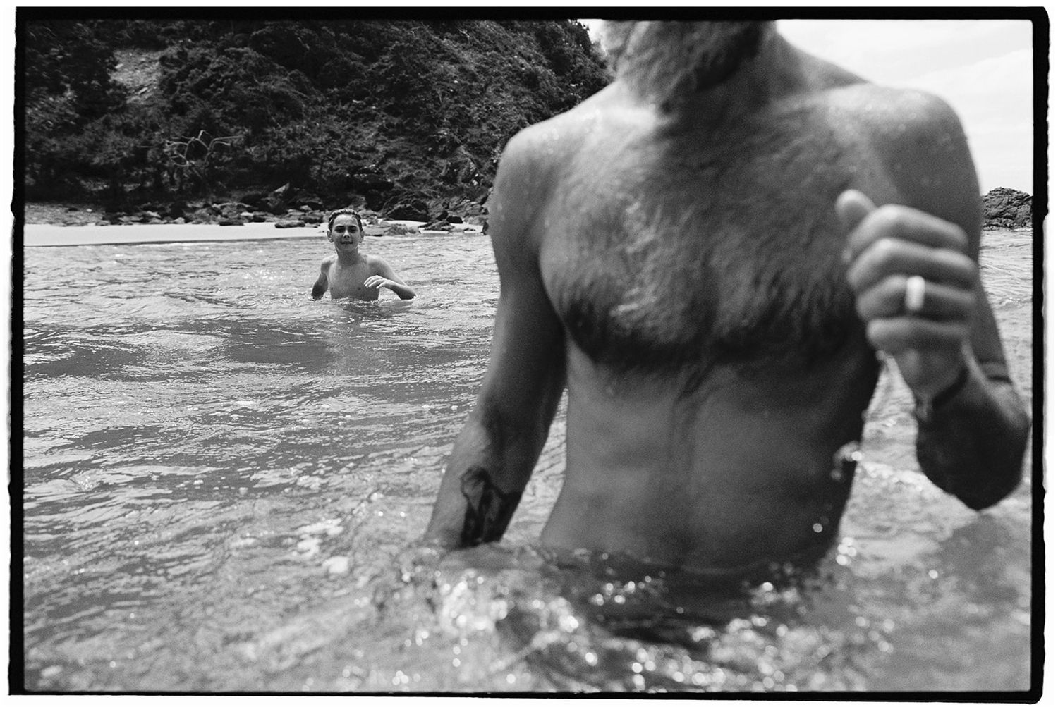 Black and white photo of two people swimming in ocean waves near a rocky shore.