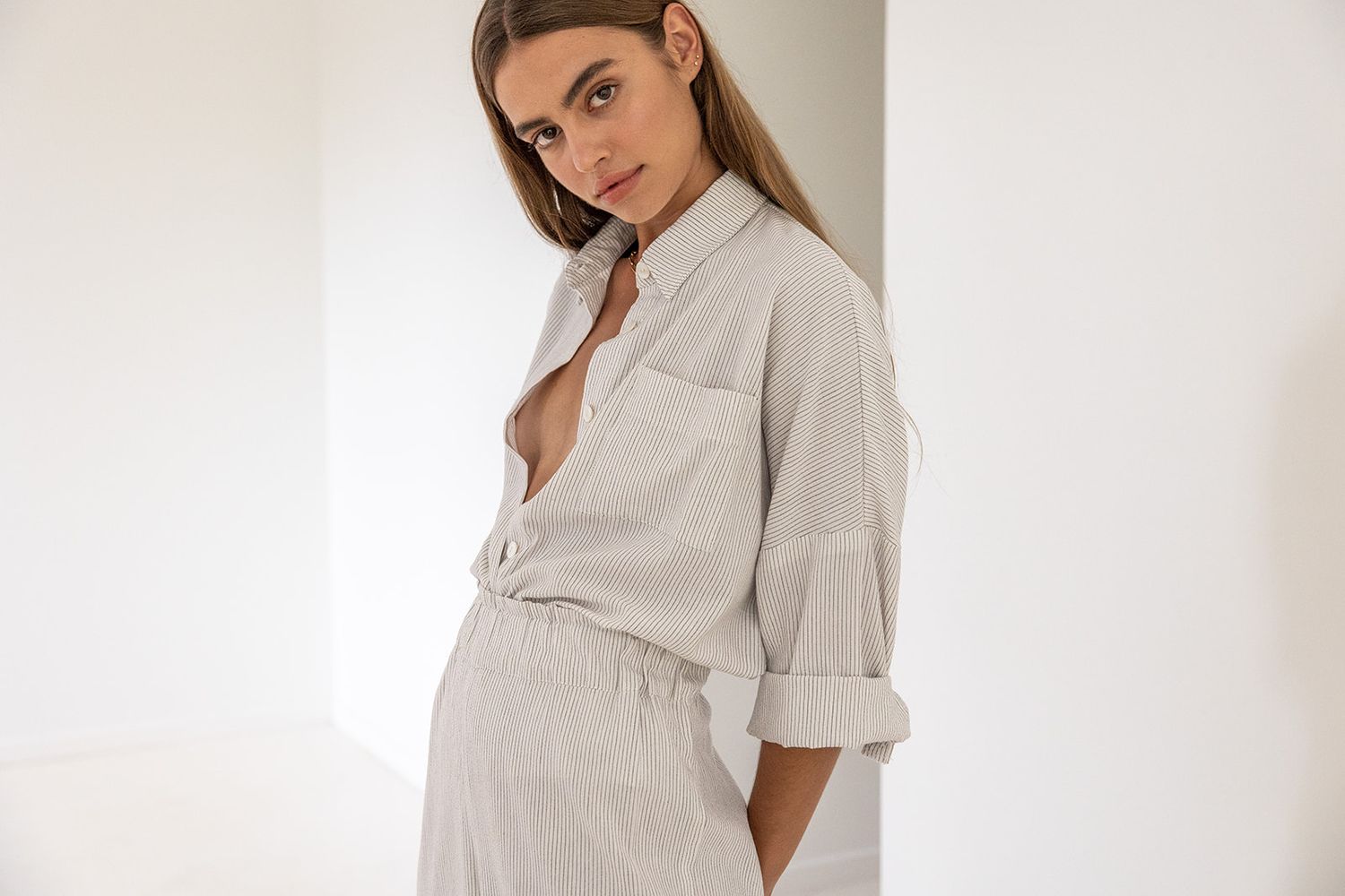 Woman in white striped oversized shirt and matching pants poses against white wall in minimalist setting.