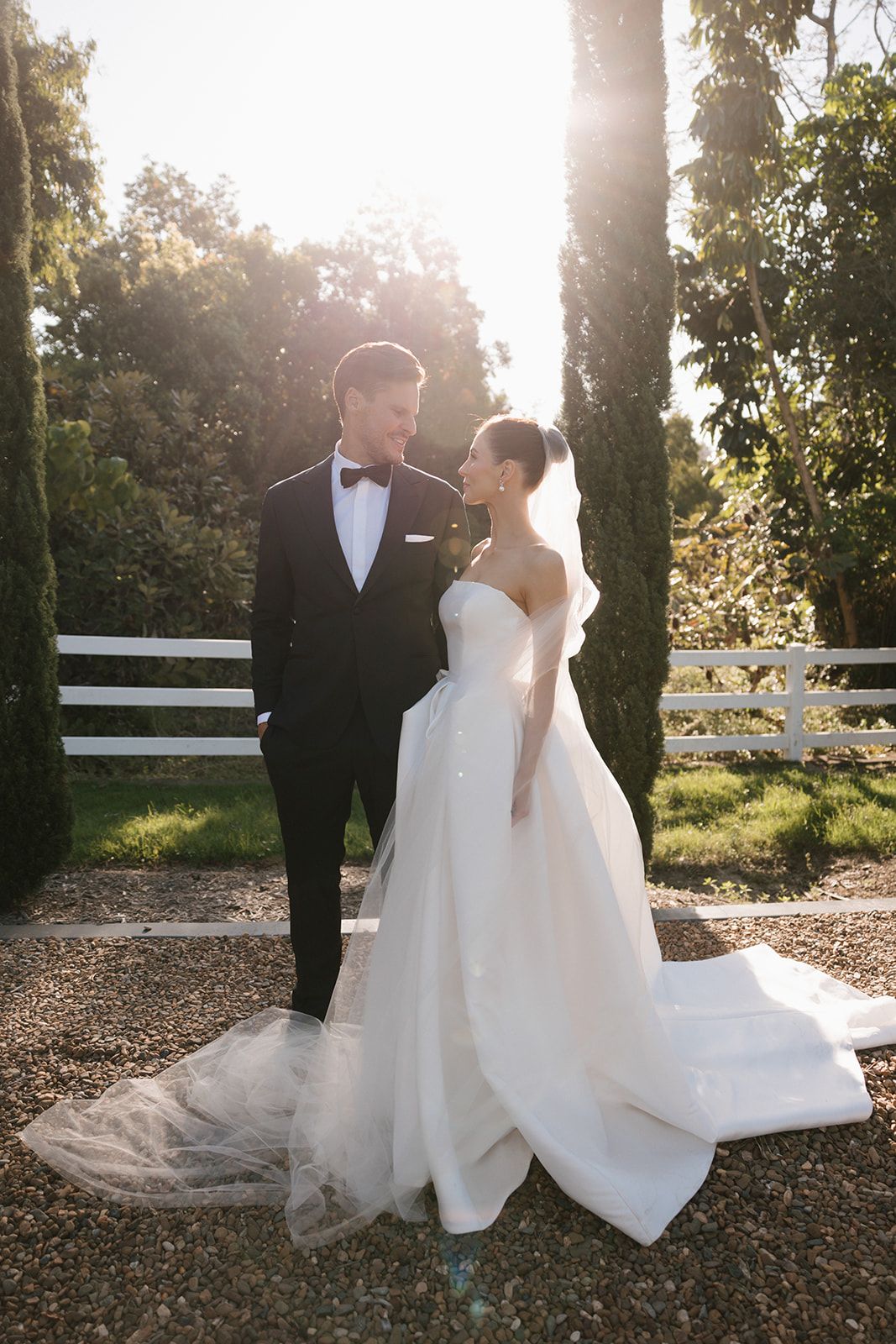 A couple in formal wedding attire poses together at sunset near white fencing and trees at an outdoor venue.