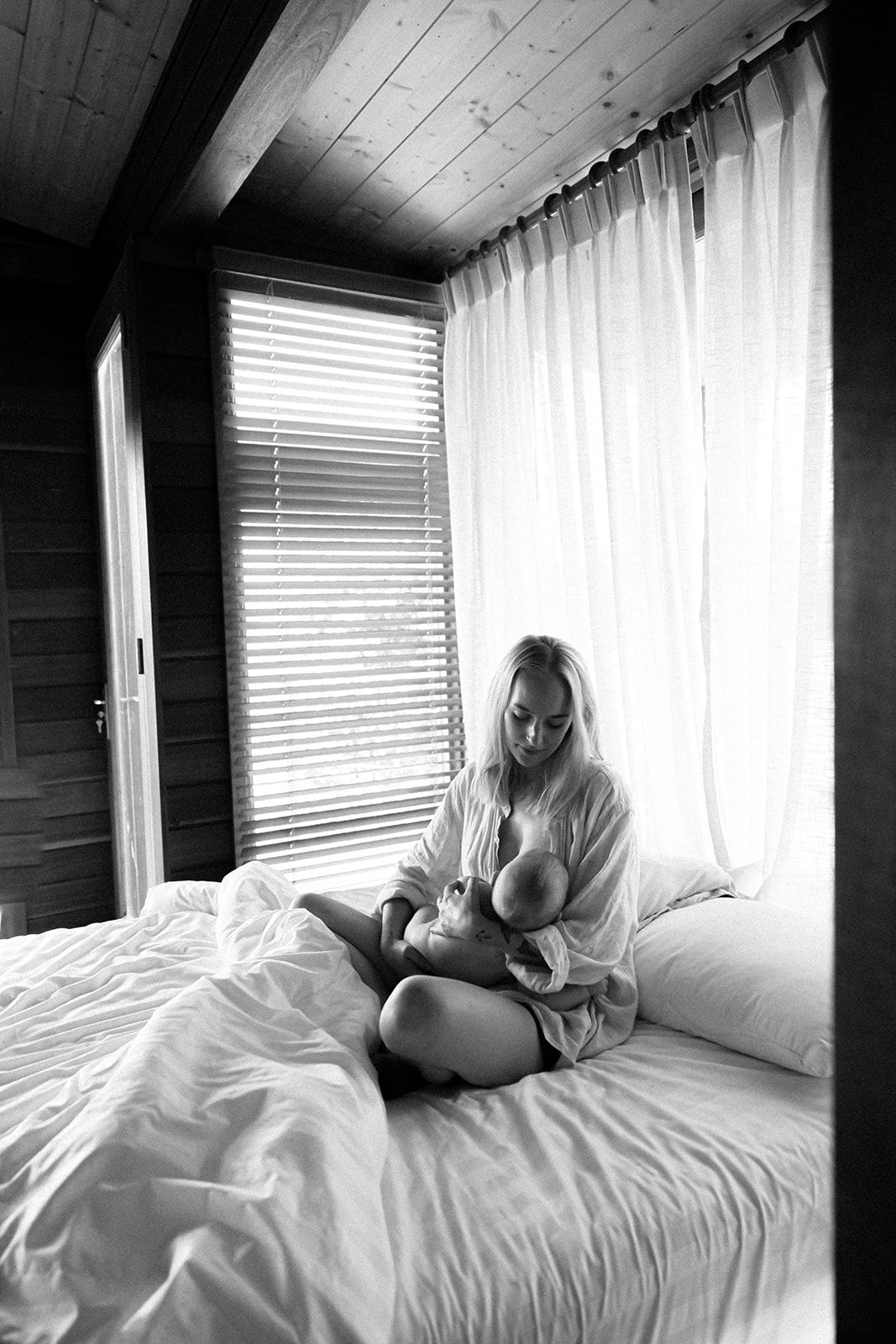 Black and white photo of someone sitting cross-legged on a bed by a window with blinds and sheer curtains.