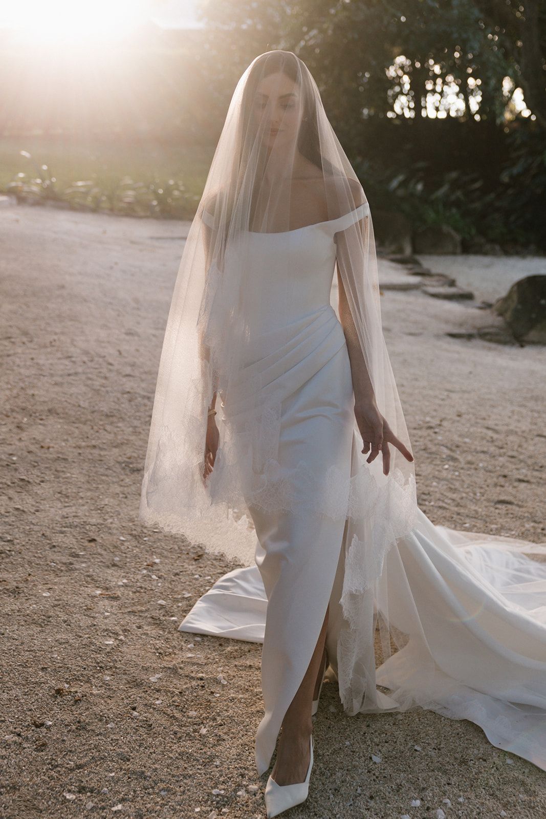 Bride in an off-the-shoulder satin wedding gown with a thigh-high slit, sheer veil and long train, walking in warm golden sunlight.