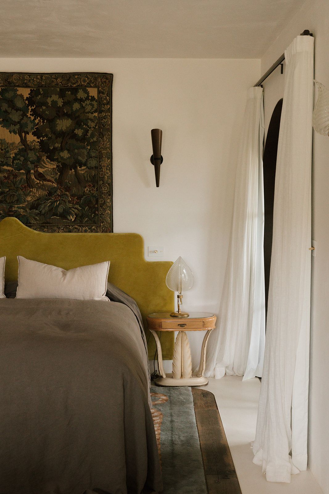 Elegant bedroom with olive velvet headboard, tapestry wall art, sheer white curtains, and vintage bedside lamp.
