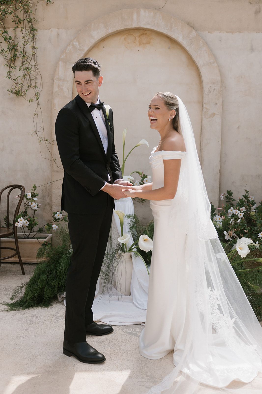 A bride and groom share a joyful moment during their wedding ceremony against a light stone wall backdrop.
