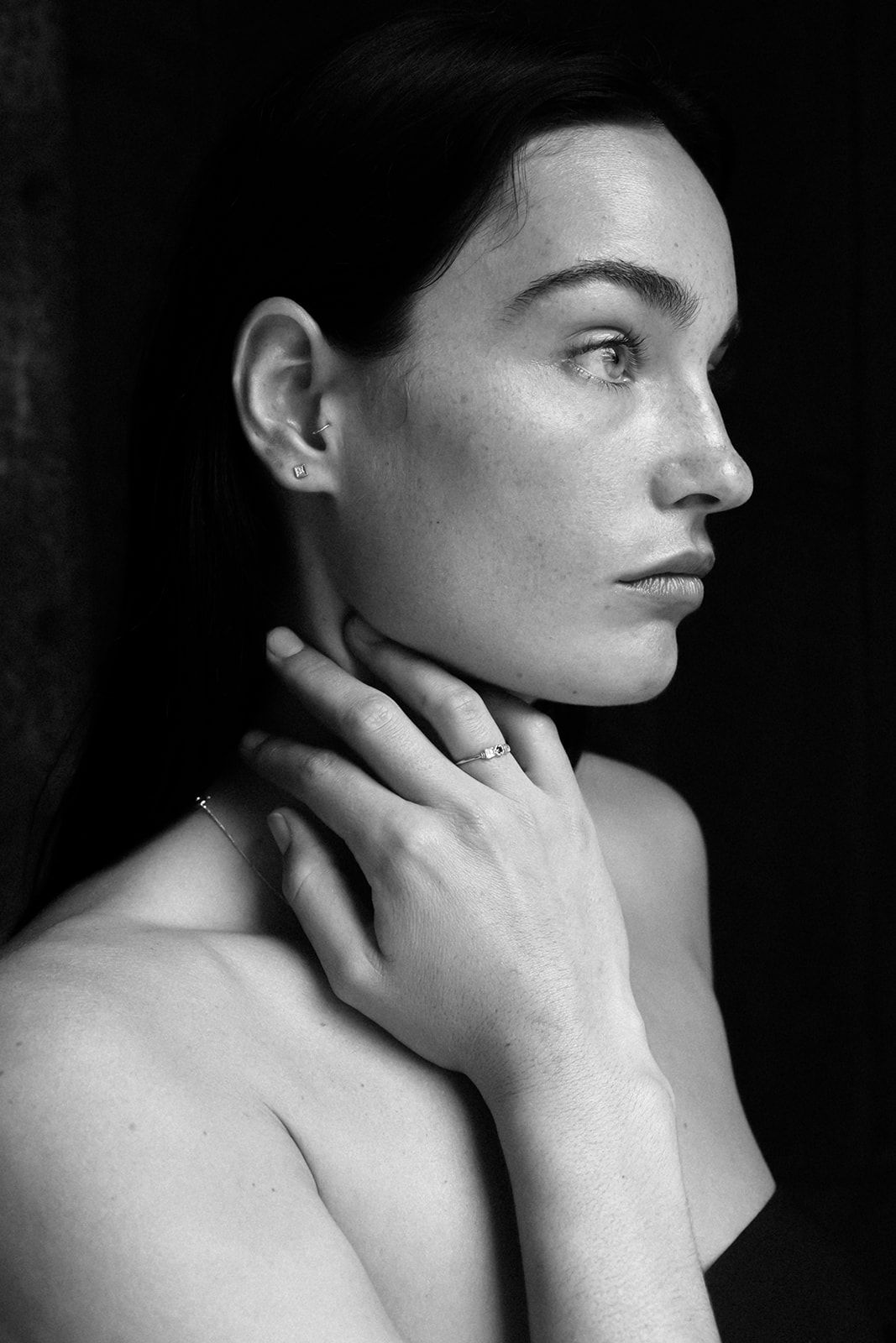 Black and white portrait of a young woman with dark hair, hand on neck, wearing a delicate ring, against dark background.