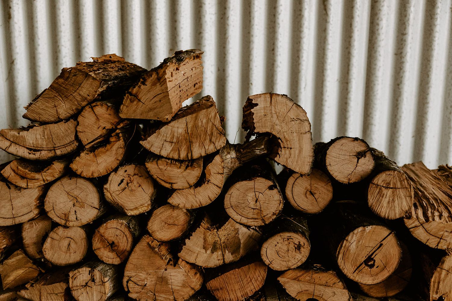 Stacked split firewood logs piled against a corrugated metal wall, showing natural wood grain and tree rings.
