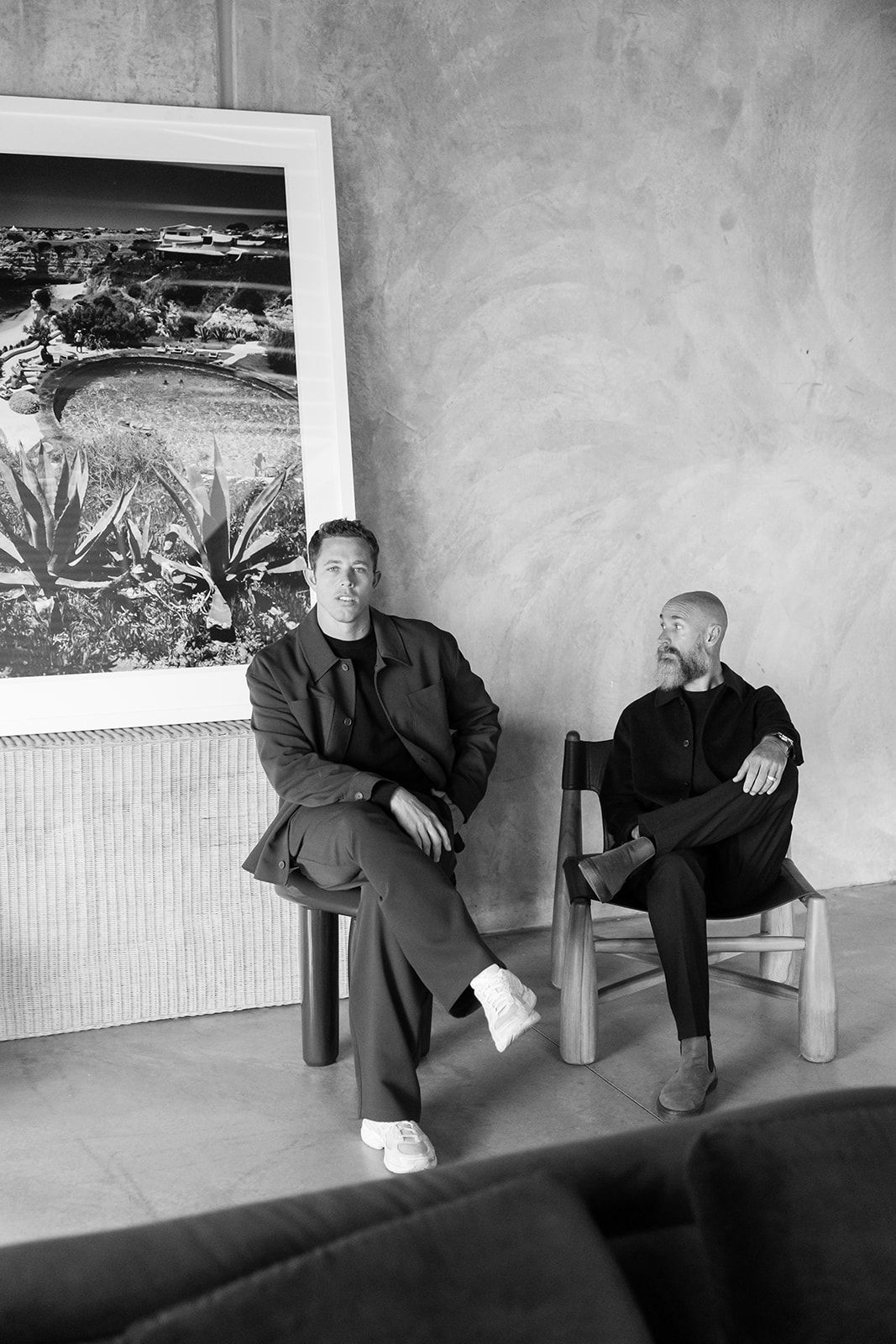 Two people sit casually in chairs against a textured wall with a large framed landscape artwork displayed above them.