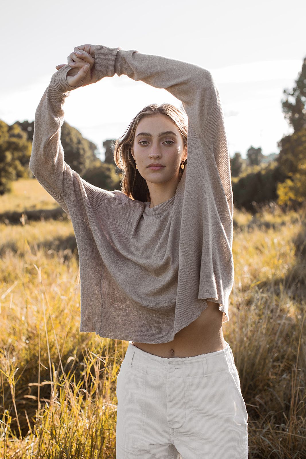 Person in gray sweater and white pants poses in golden field at sunset with arms raised.