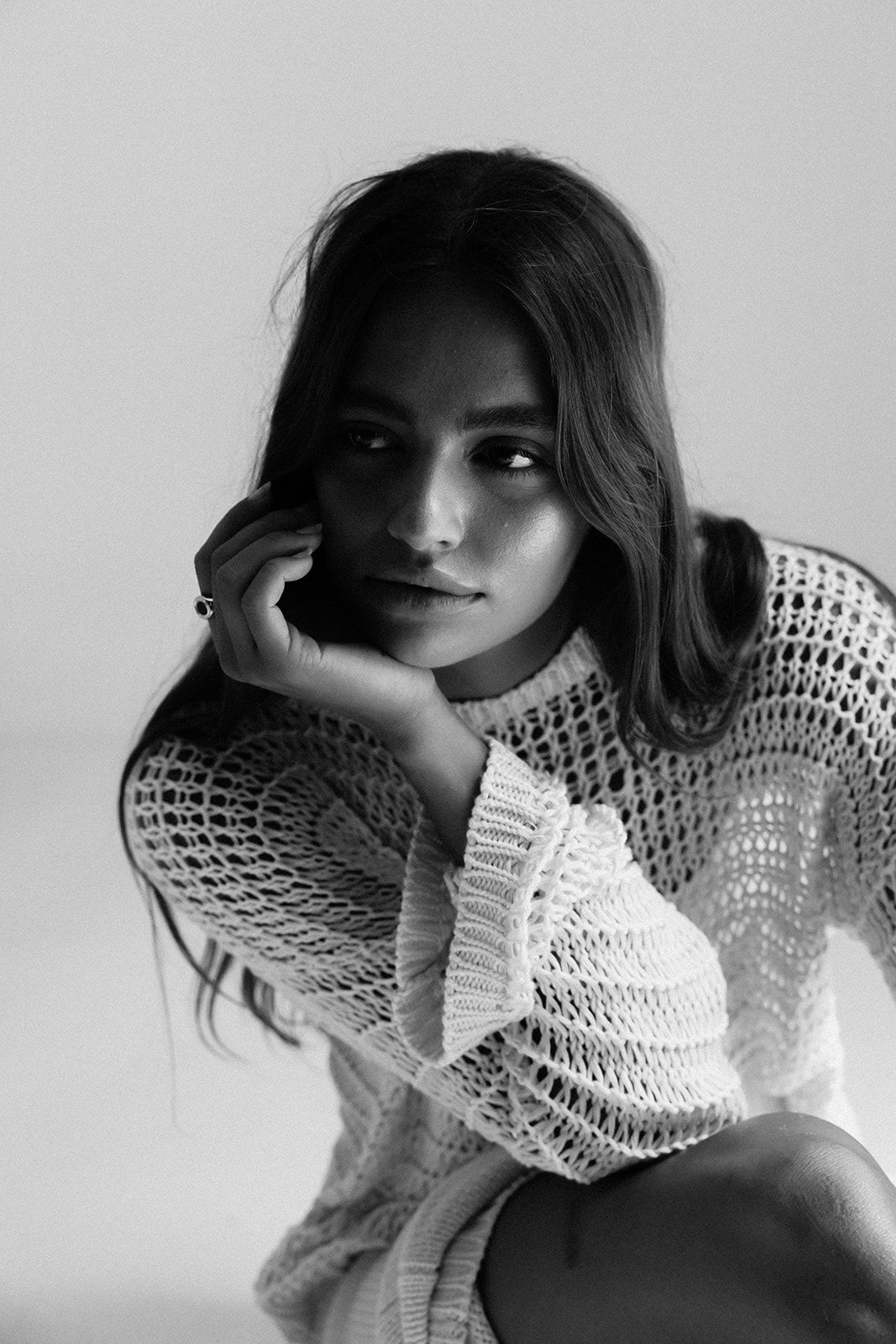 Black and white portrait of a young woman in a crochet sweater, resting her chin on her hand thoughtfully.