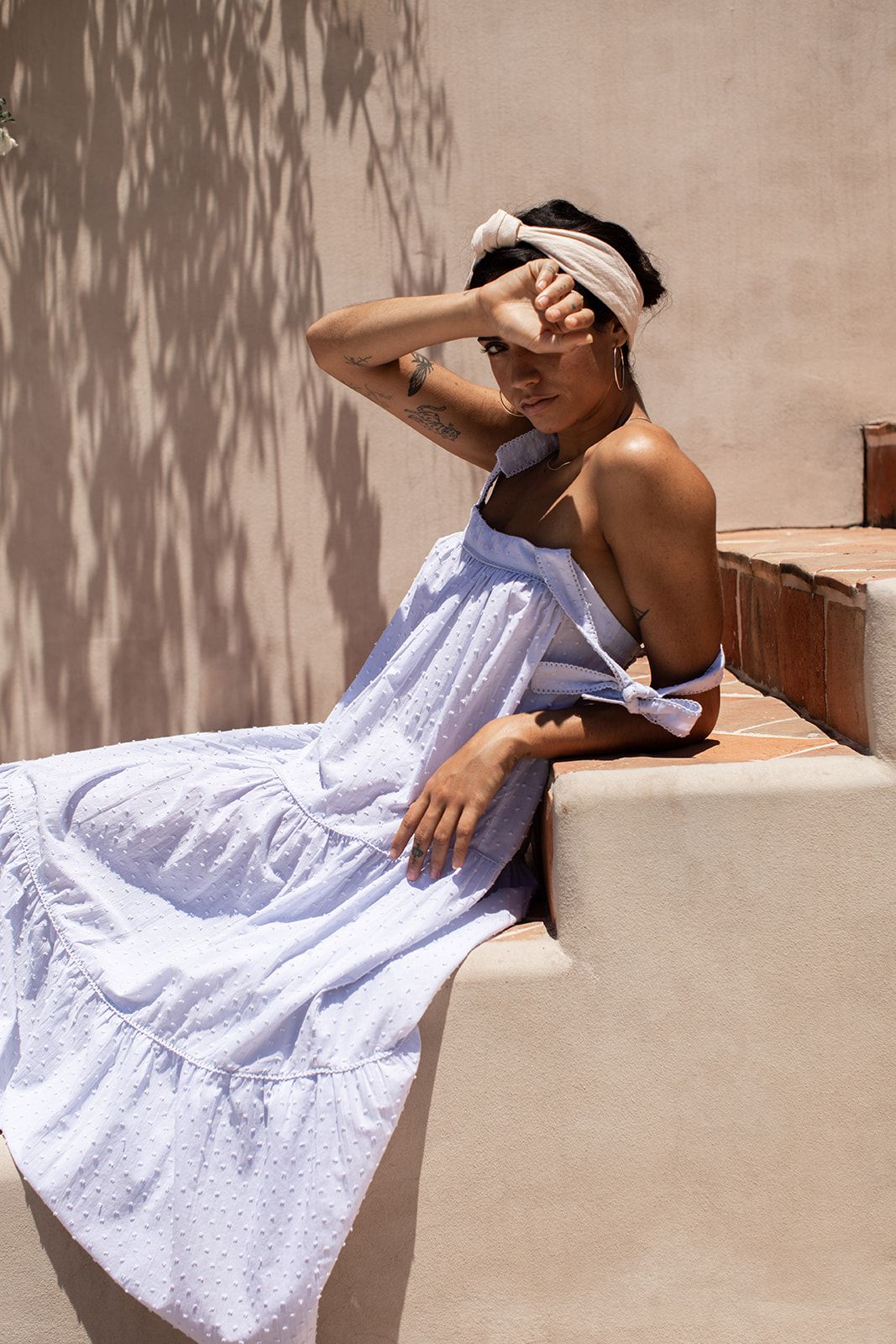A person in a white sundress and headscarf lounges on stone steps in dappled sunlight against a beige wall.
