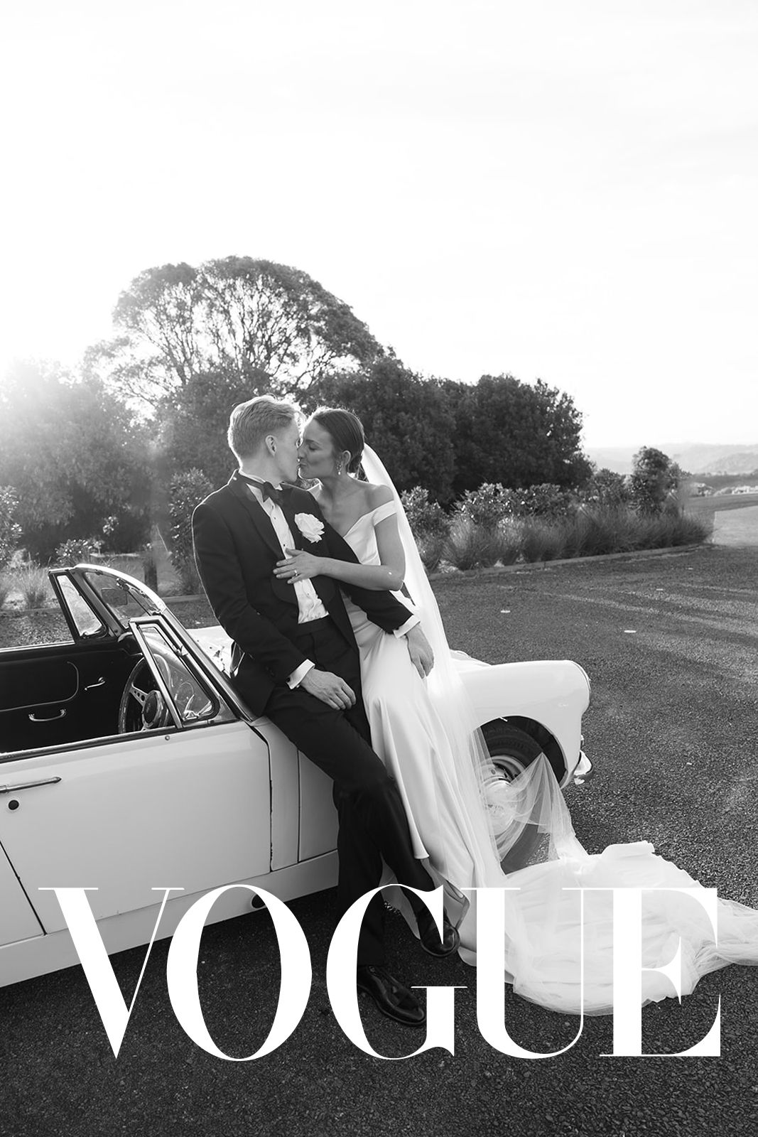 Black and white Vogue wedding photo of bride and groom kissing beside a vintage convertible car outdoors.
