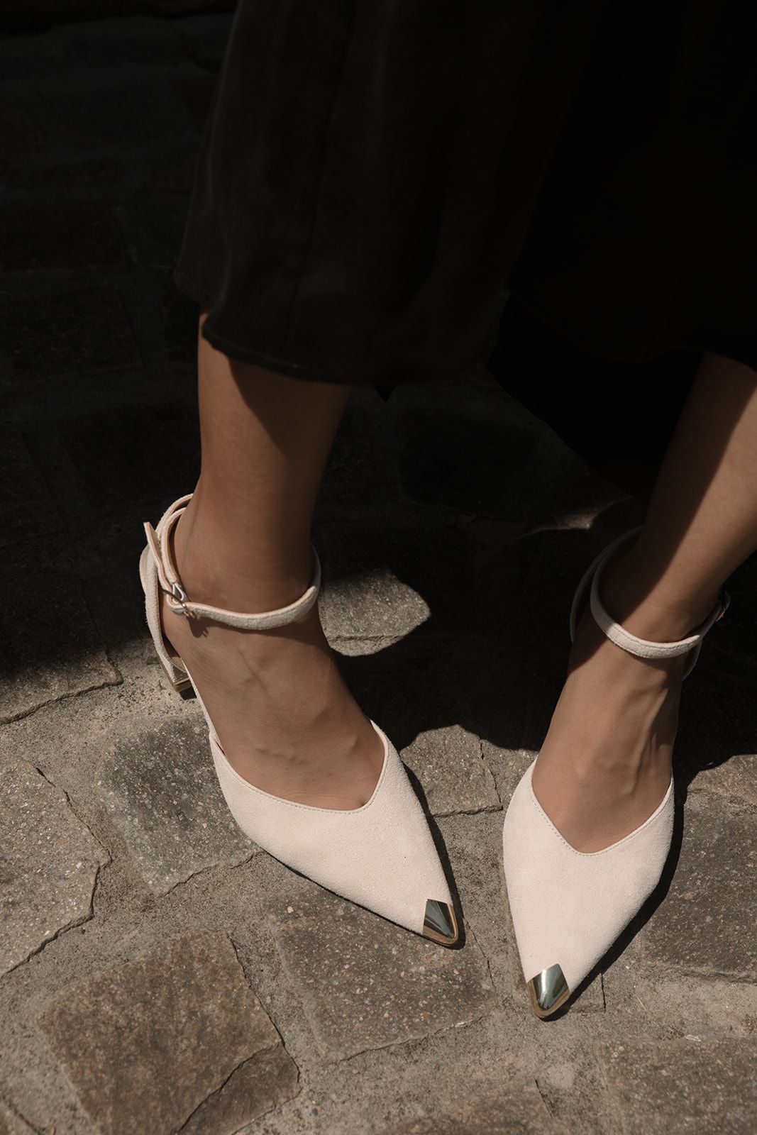 Elegant white pointed-toe shoes with metal tips and ankle straps shown against dark stone flooring.