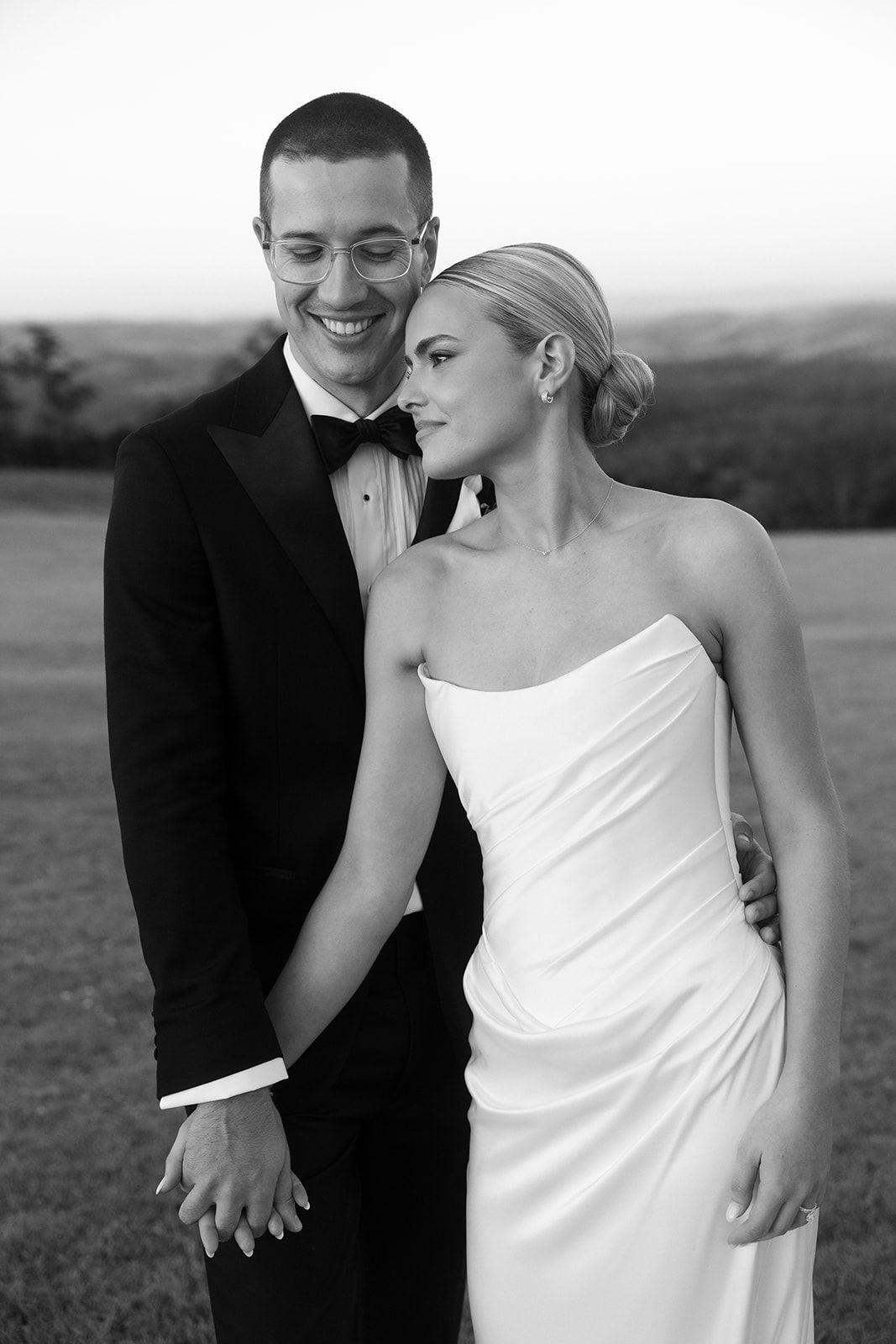 Black and white wedding portrait of smiling groom in tuxedo and bride in strapless gown holding hands outdoors.