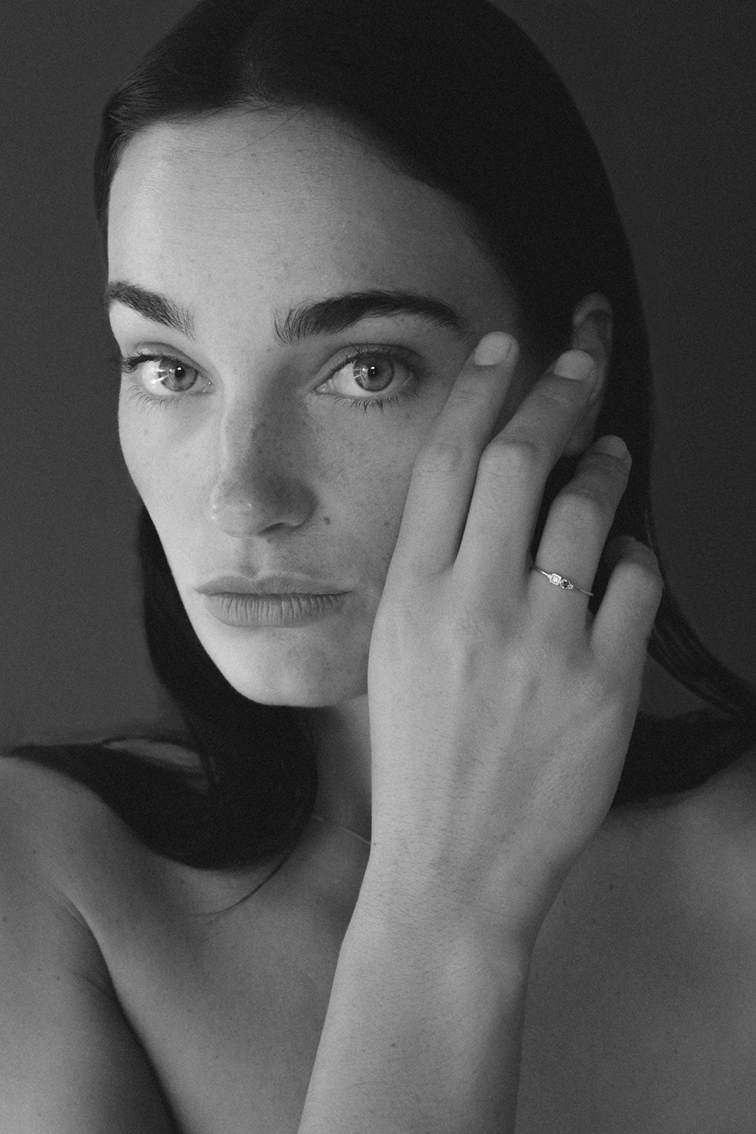 Black and white portrait of a young woman with freckles, touching her face, wearing a delicate ring.