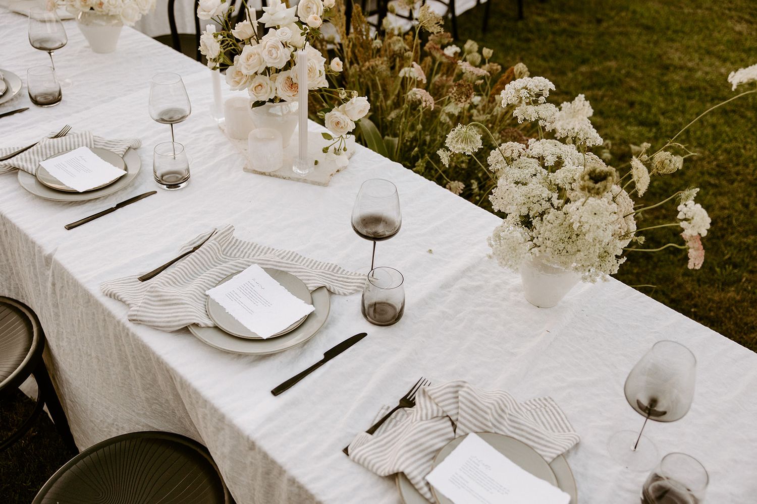 An elegant outdoor wedding table setting with white linens, floral centerpieces, and stemware in a garden setting.
