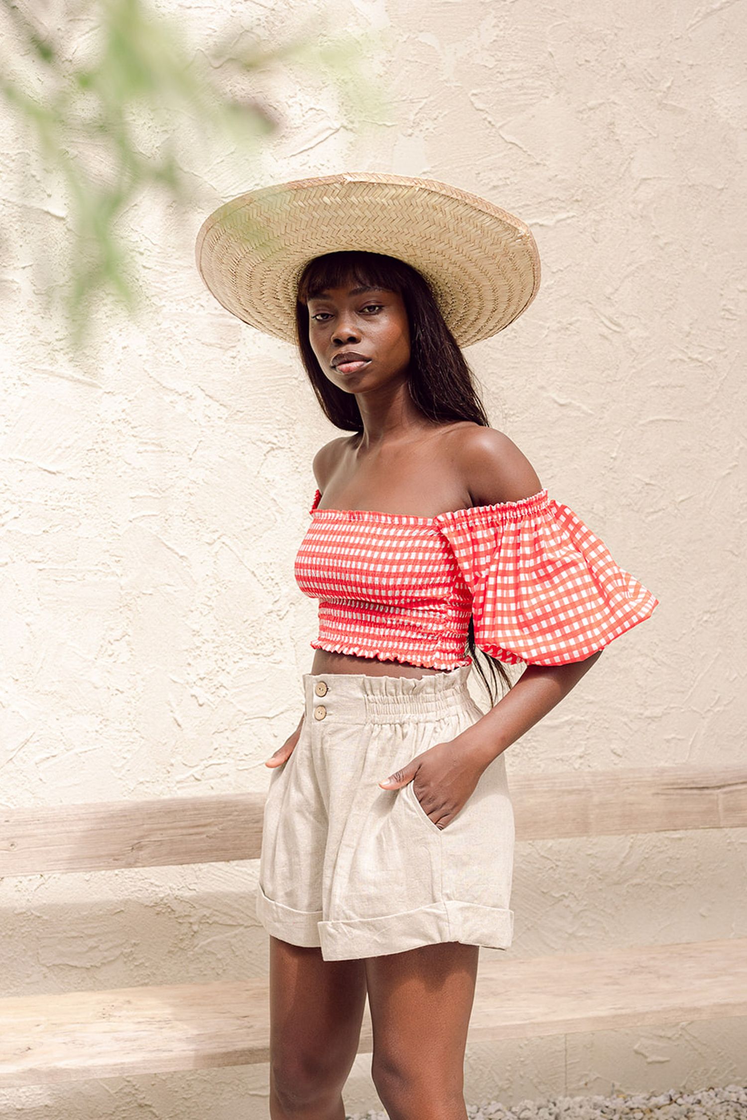 A child in a red gingham off-shoulder top and white shorts poses outdoors wearing a wide-brimmed sun hat.