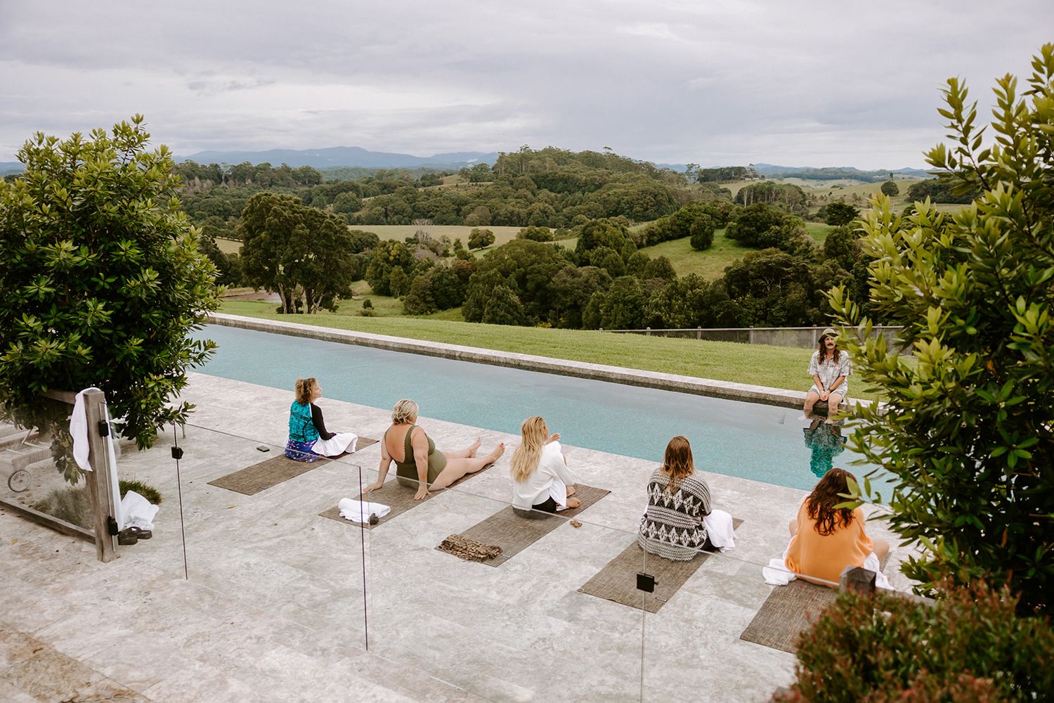 Women practicing yoga on mats beside a serene infinity pool overlooking lush green countryside landscape.
