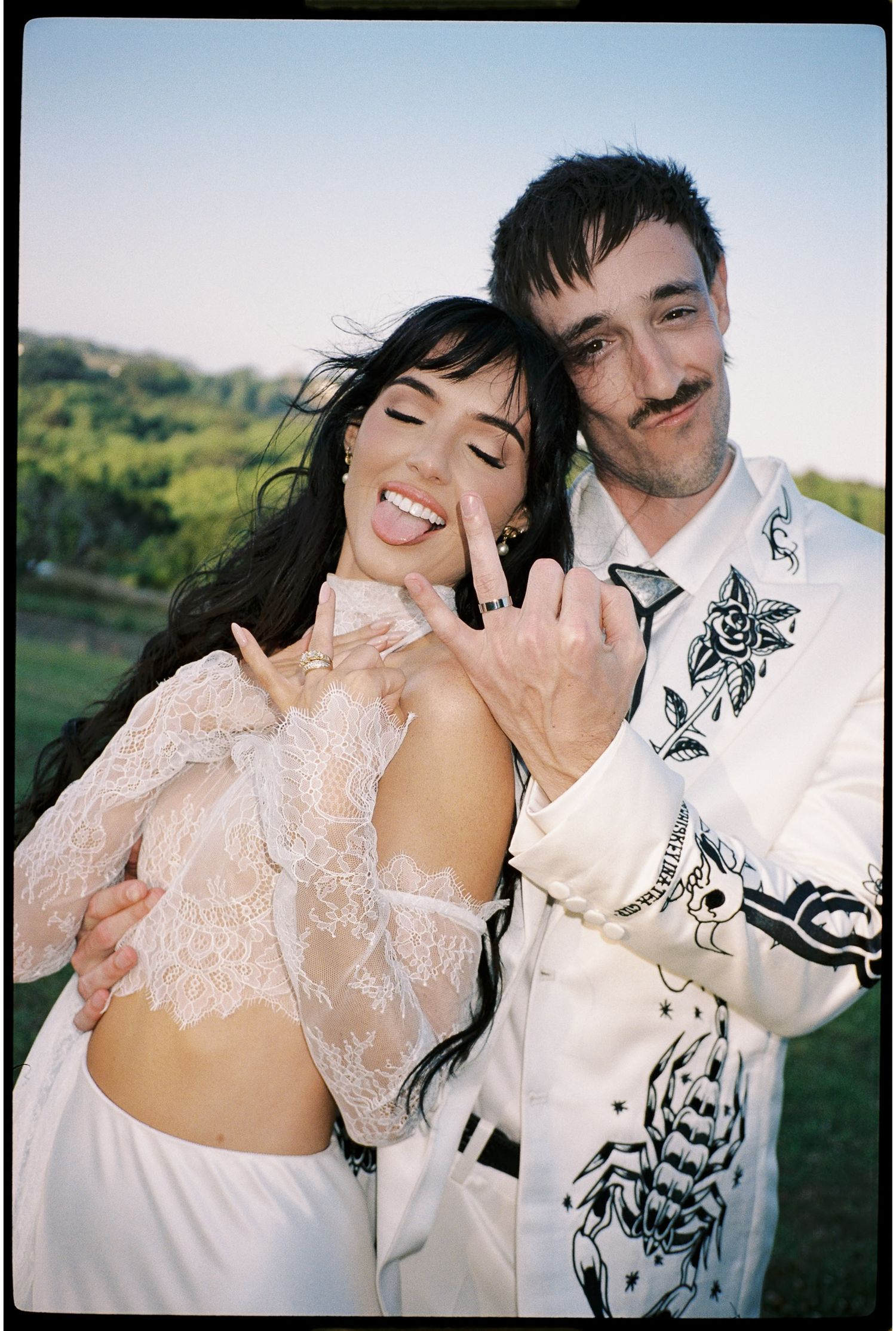 Couple in white outfits posing outdoors, woman in lace top and man in embroidered jacket, both making hand gestures.