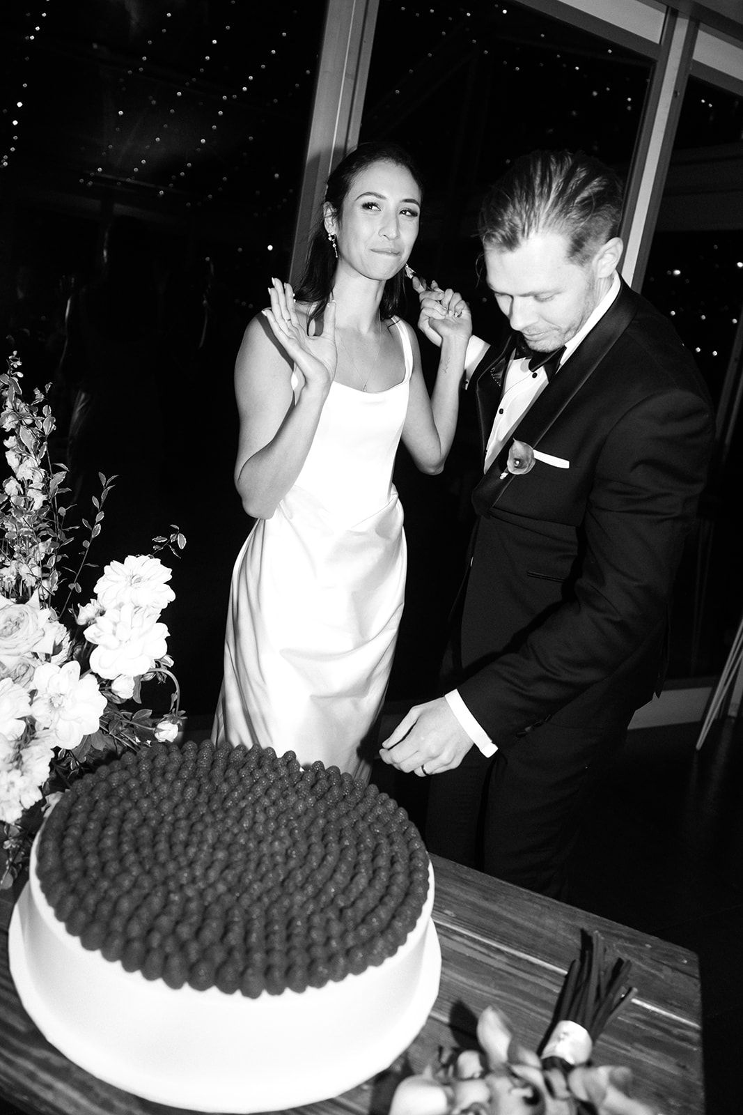 A couple in formal wedding attire looks at a large cake with dark frosting details at an evening celebration.